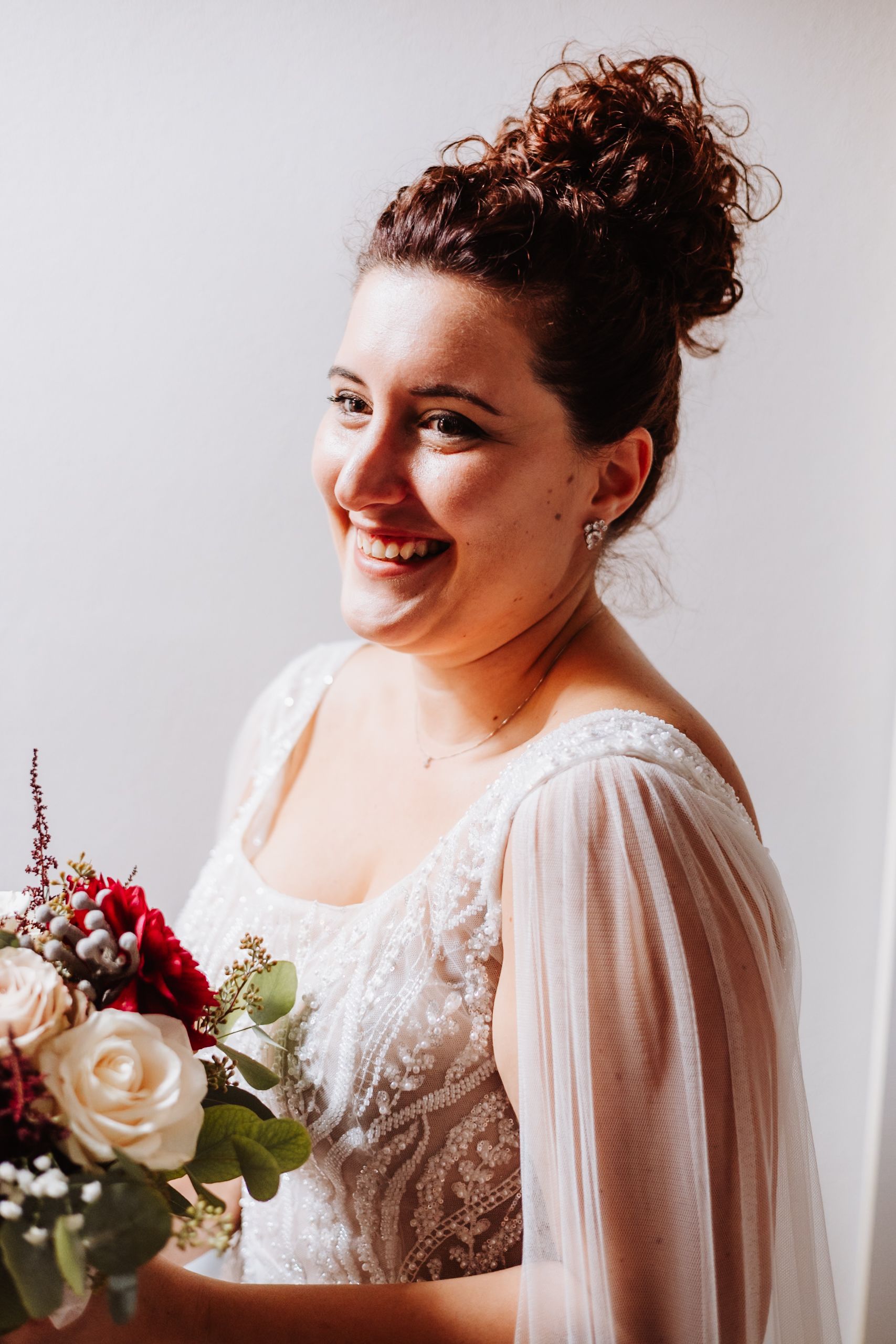 Smiling bride in white lace gown with sheer sleeves holds elegant bouquet featuring red and cream roses.