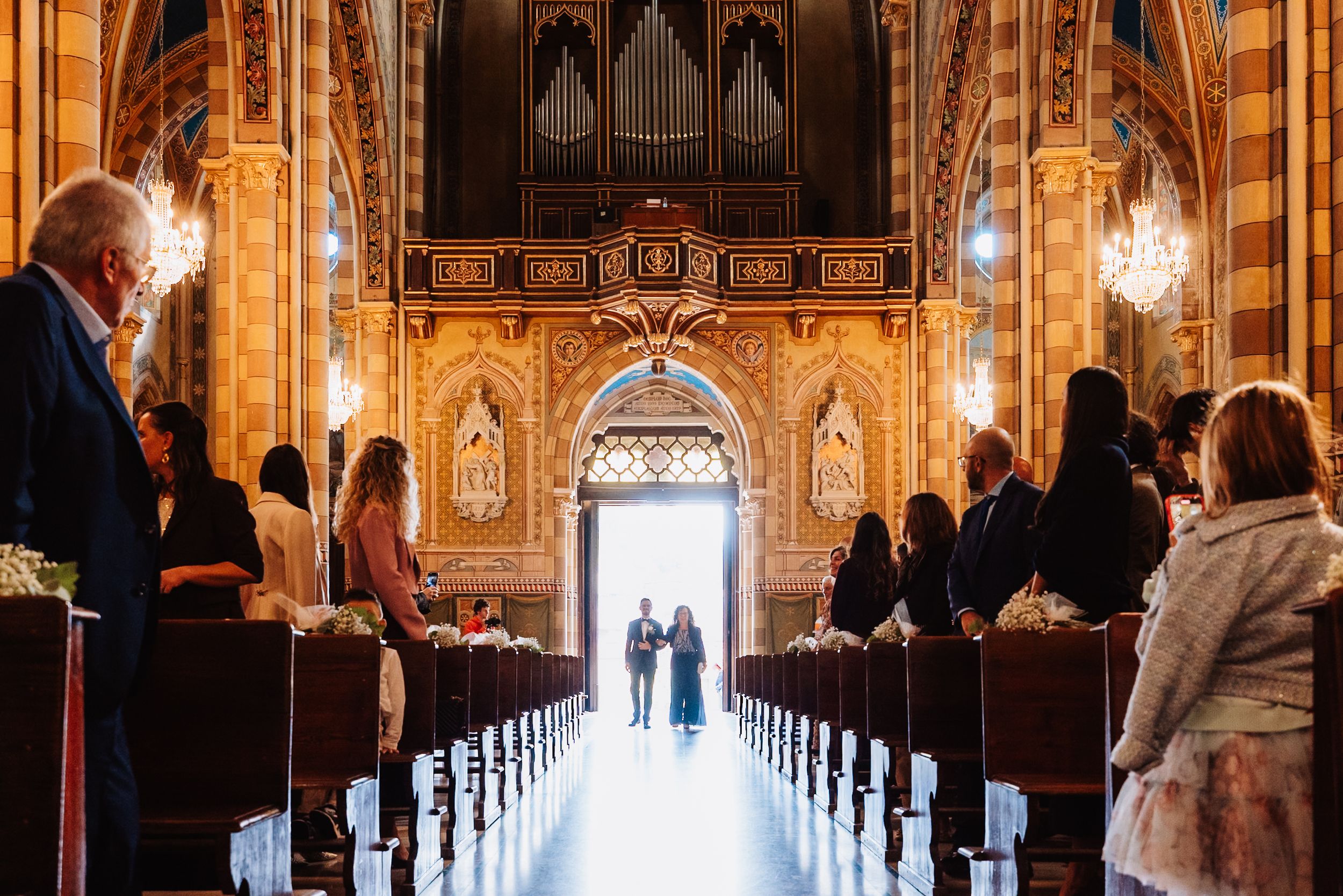 Interior view of an ornate cathedral with rows of wooden pews leading to a grand altar and stained glass windows.
