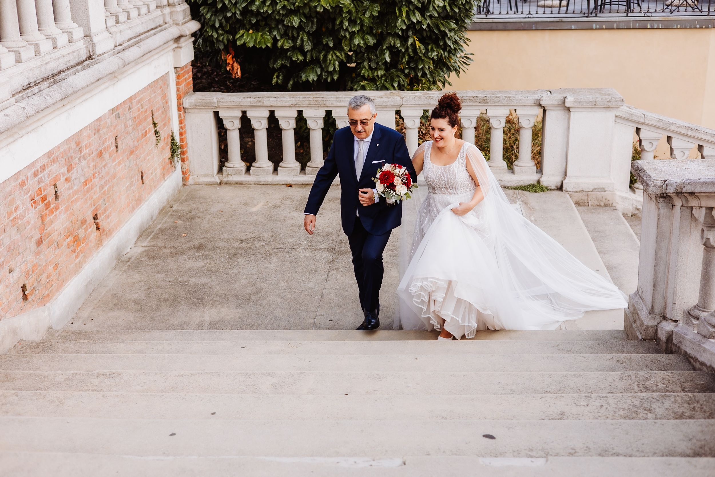 A couple in wedding attire walks up elegant stone steps at a historic venue with ornate balustrades.