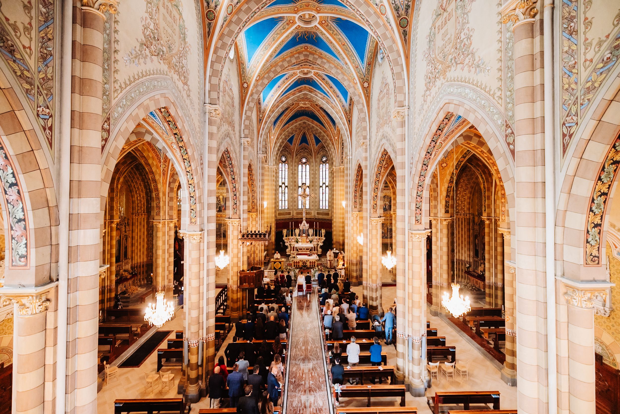 Aerial view of an ornate Gothic cathedral interior with vaulted blue and gold ceilings and people seated in wooden pews.
