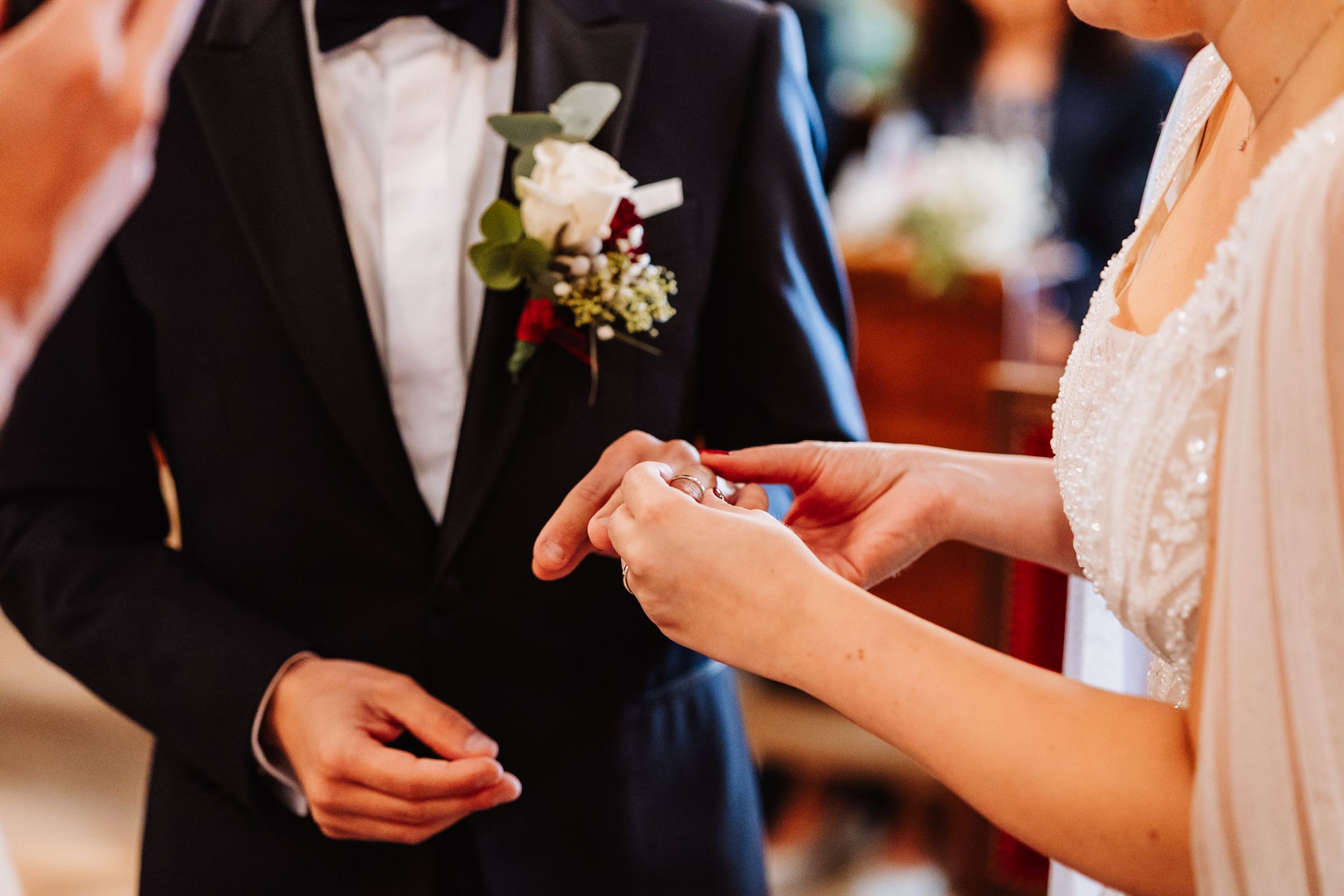 Close-up of hands exchanging wedding rings during ceremony with groom wearing black suit and white boutonniere.