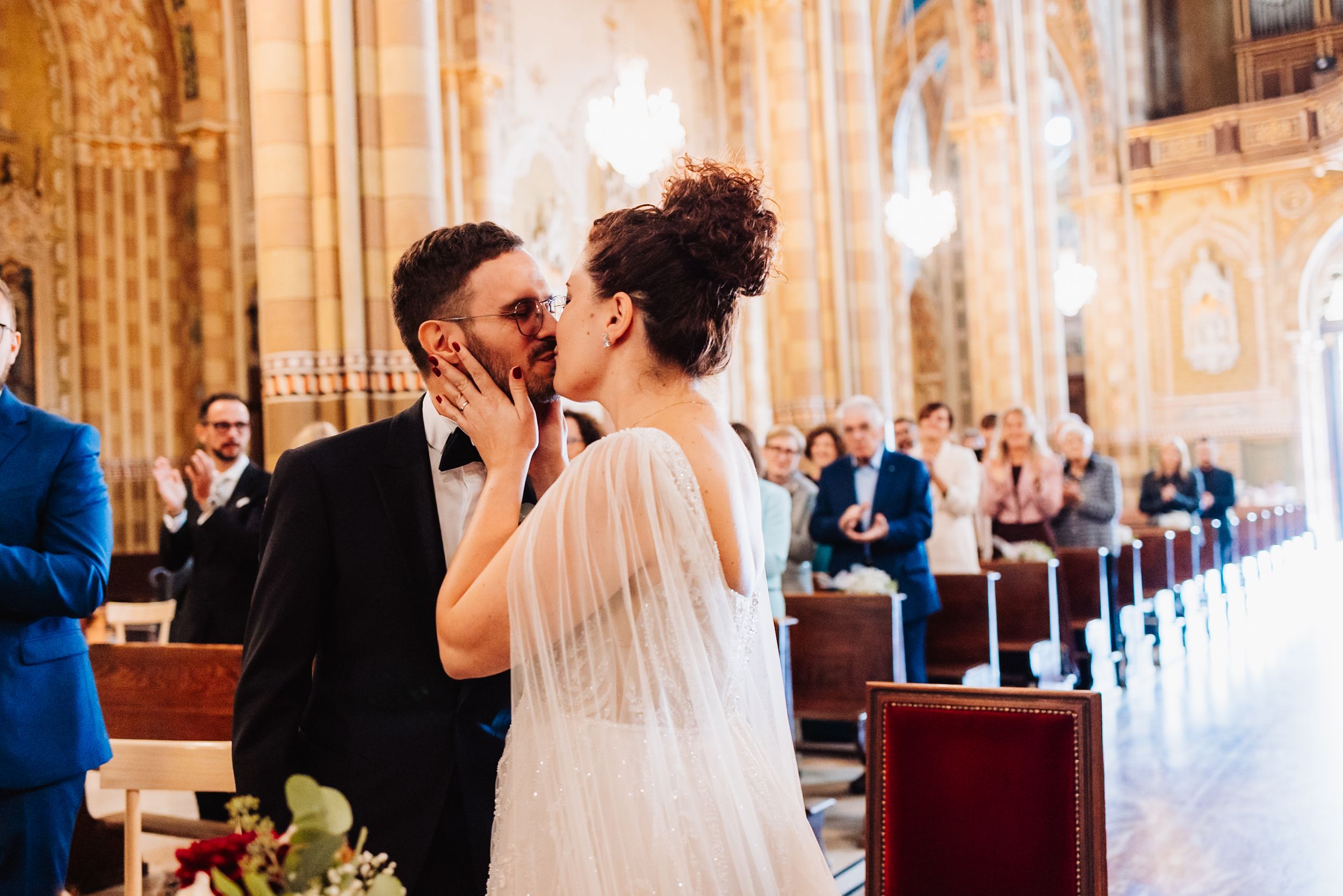 A couple shares a romantic kiss during their wedding ceremony in an ornate cathedral with golden decor and pews.
