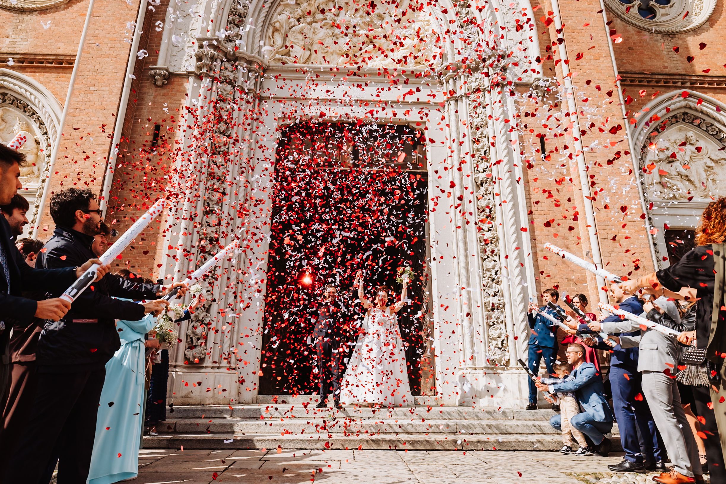 Wedding guests shower newlyweds with red confetti petals on ornate church steps during celebration.