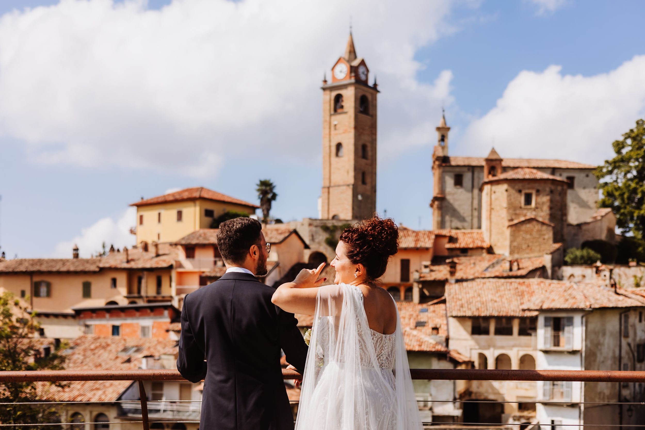 A couple takes photos of a scenic Italian hilltop town with terracotta roofs and a historic bell tower against blue skies.