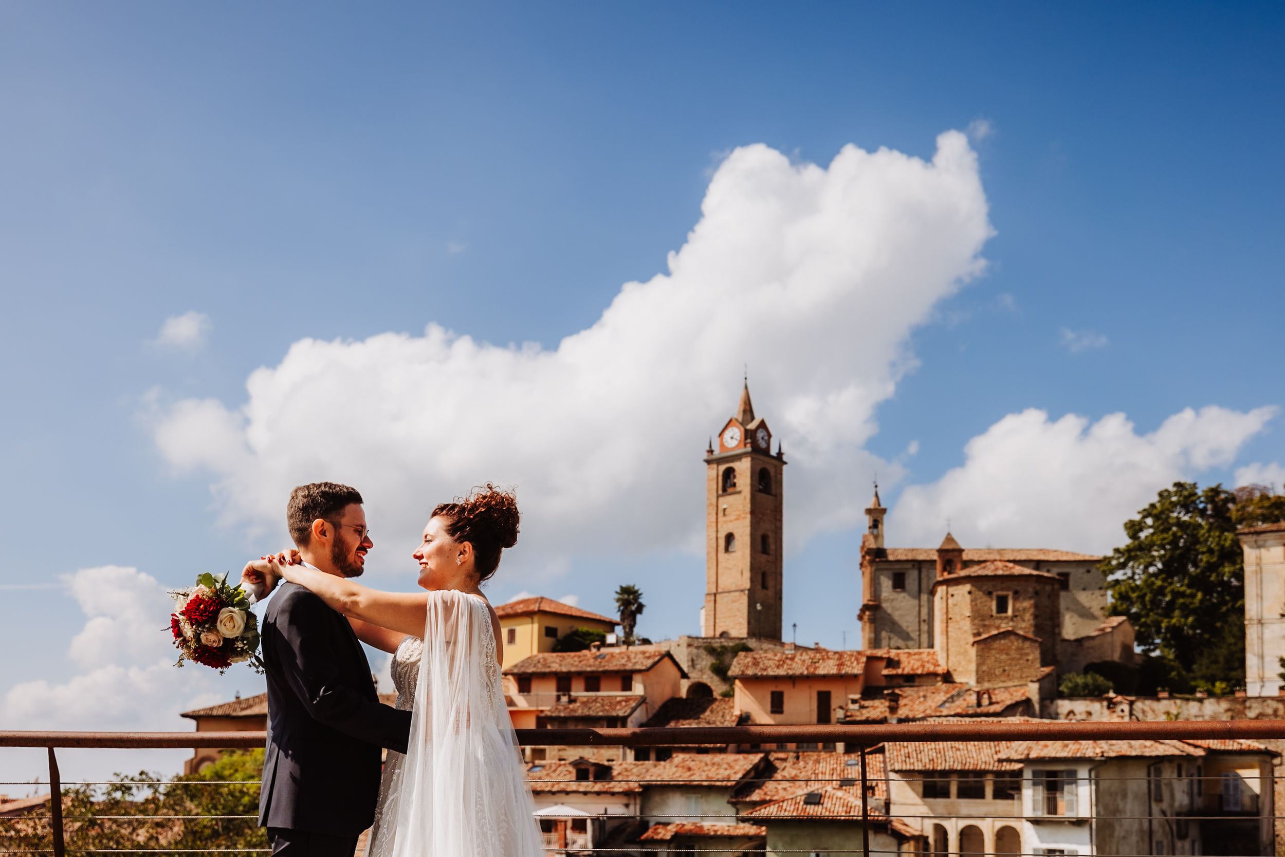 Wedding couple embraces against backdrop of historic Italian town with medieval towers and architecture under blue sky.