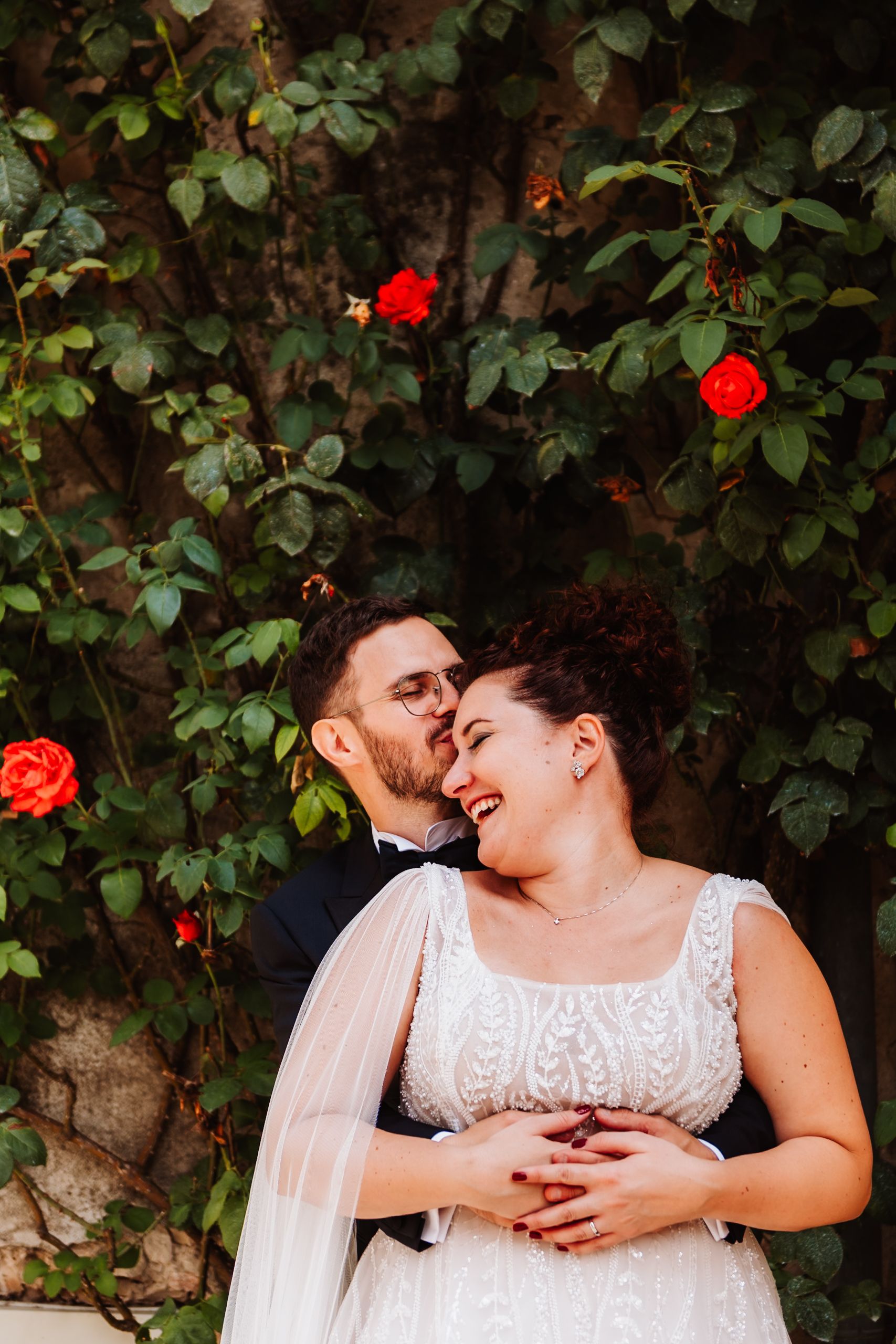 A couple in white attire share an intimate moment while laughing together against a backdrop of green foliage and red flowers.
