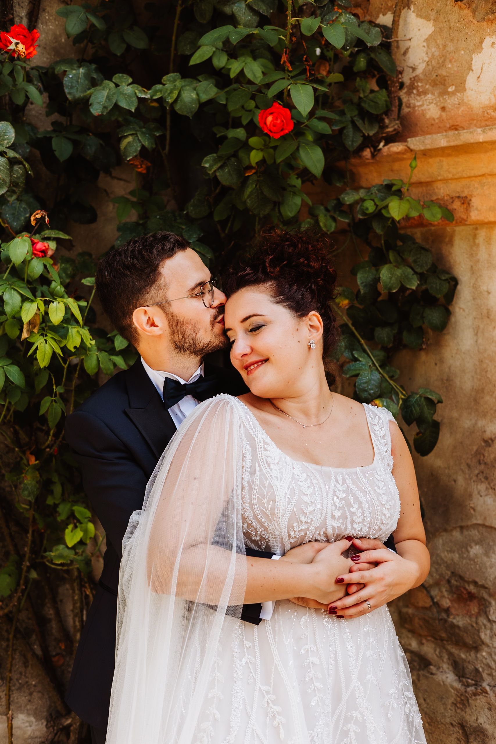 A romantic wedding couple embraces near a stone wall with red roses and green foliage in the background.