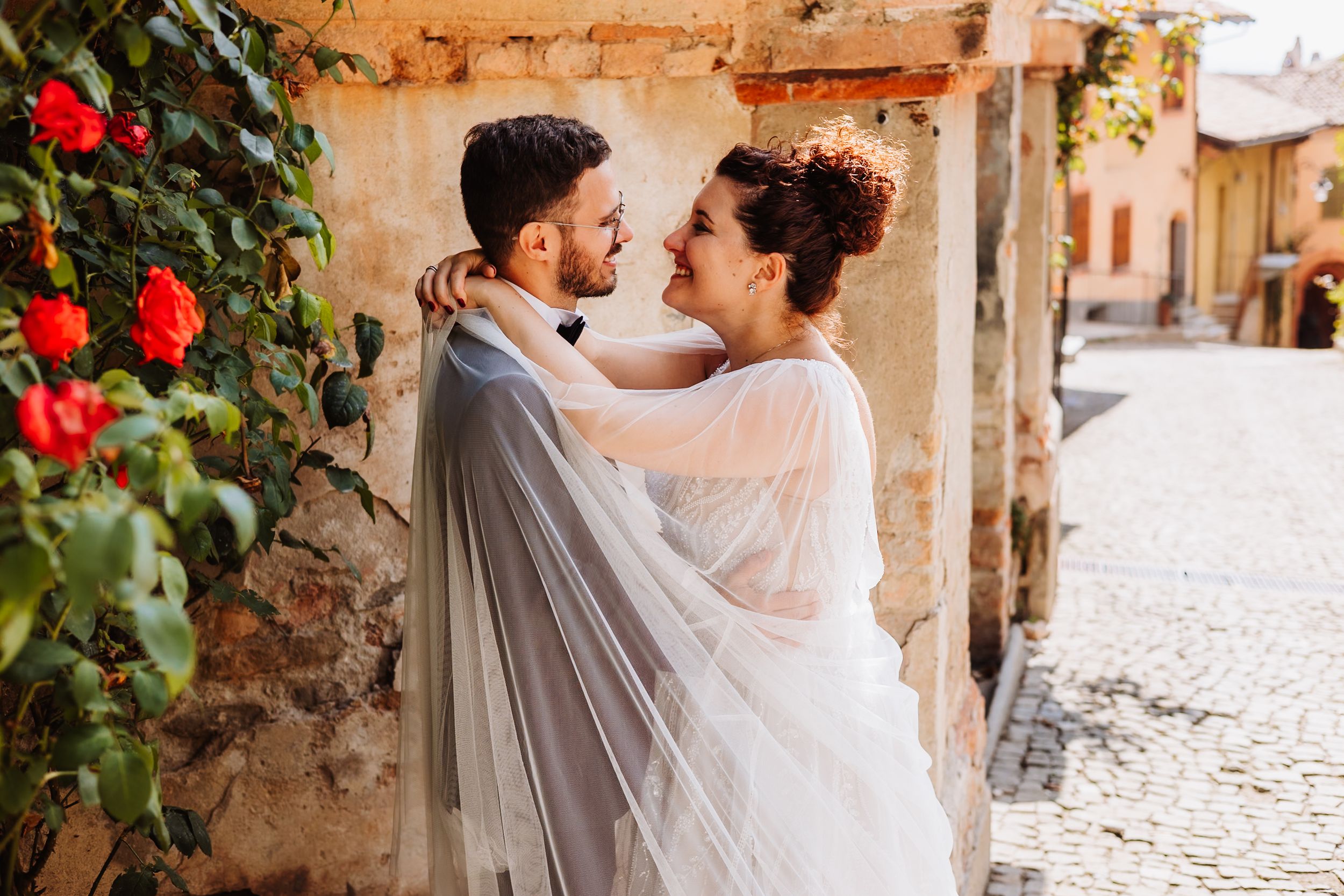 Couple embraces in a romantic moment along a rustic stone wall with red flowers in an Italian village setting.