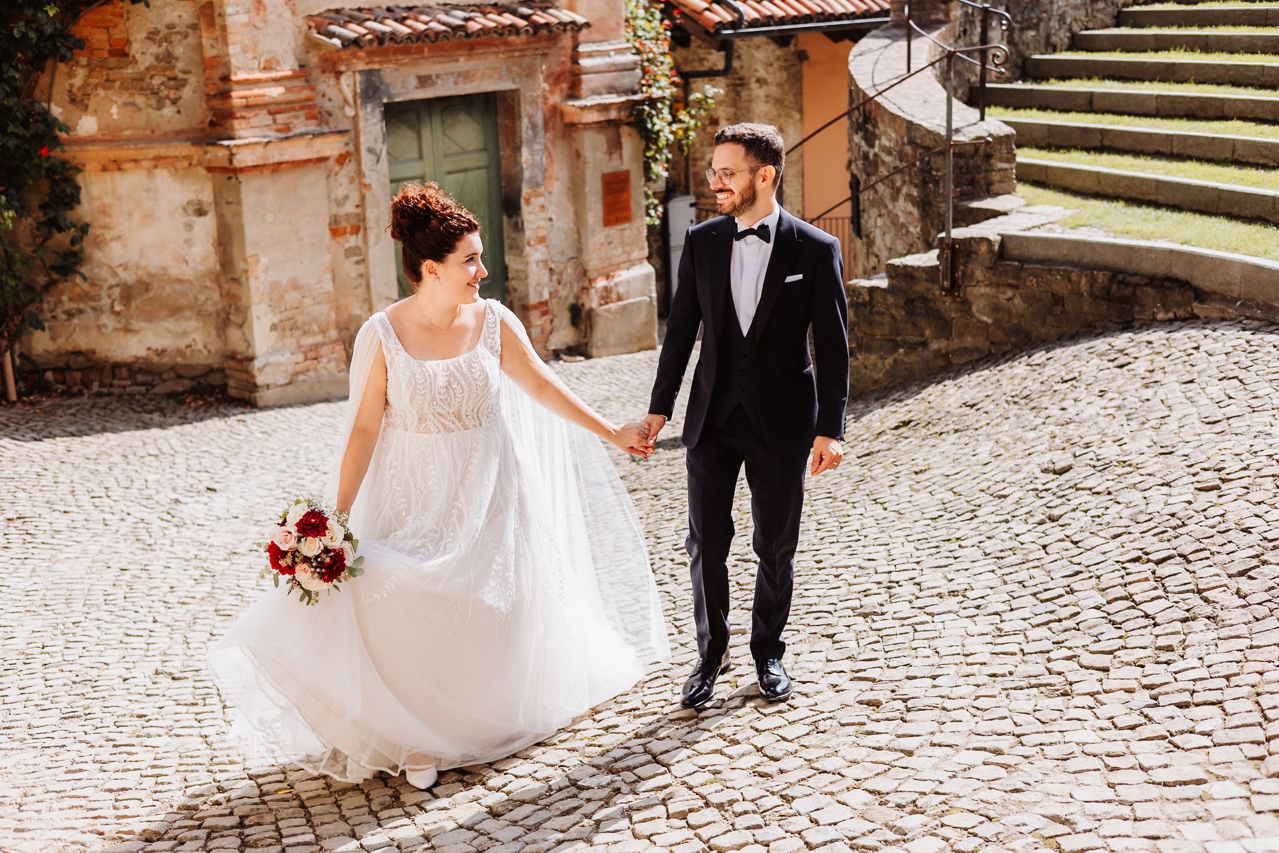 Couple in wedding attire walking hand in hand on a charming cobblestone street in a historic European town.