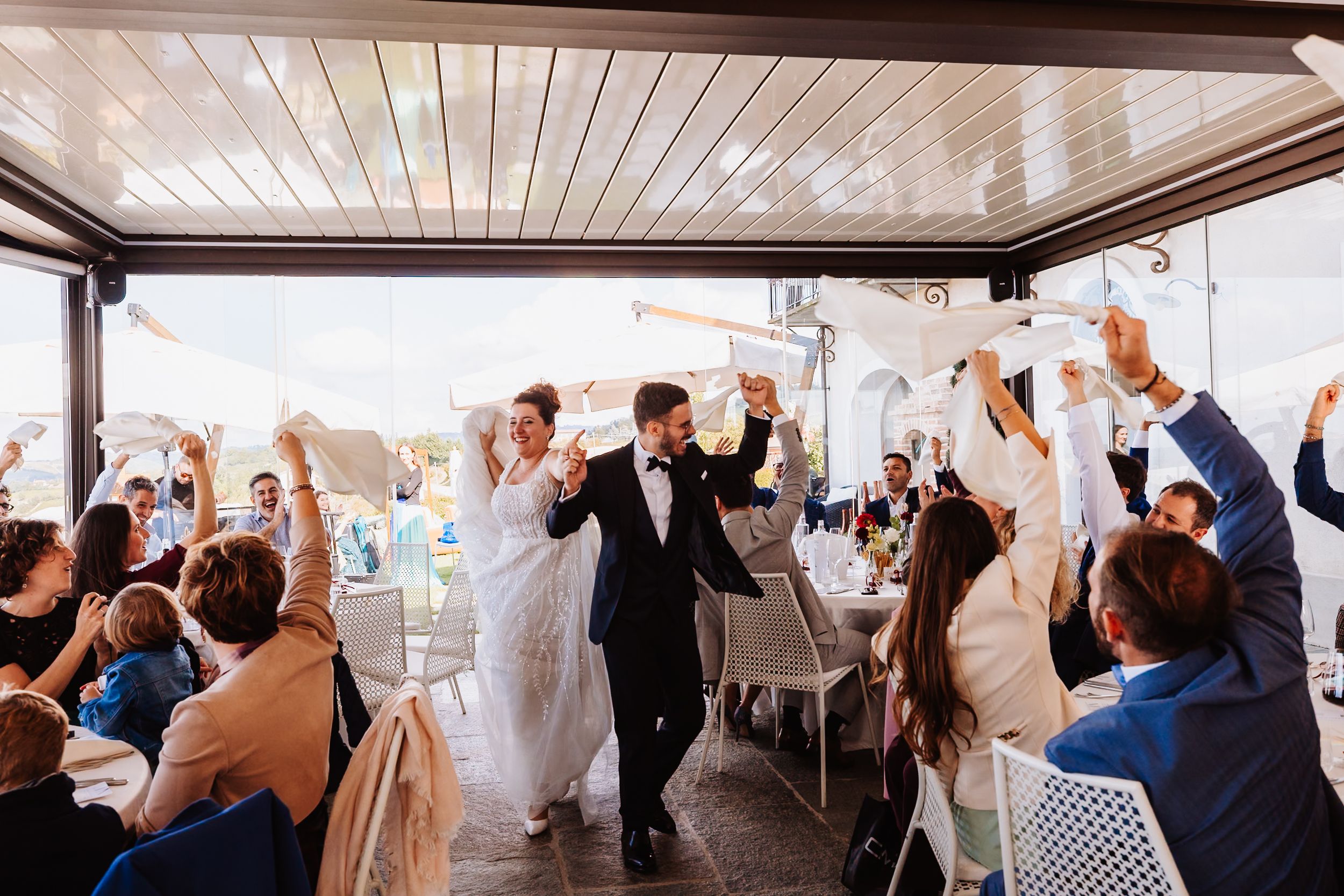 Wedding guests raise their hands in celebration during a reception in a bright, modern venue with white decor.