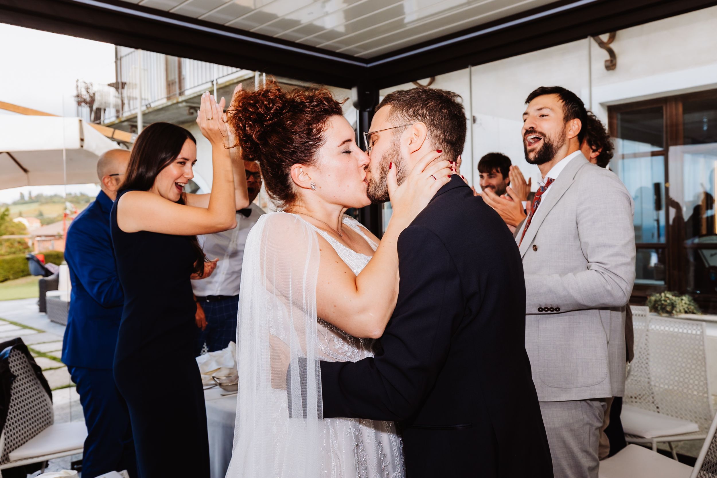 A couple shares a romantic kiss at their wedding celebration while guests cheer and celebrate in the background.