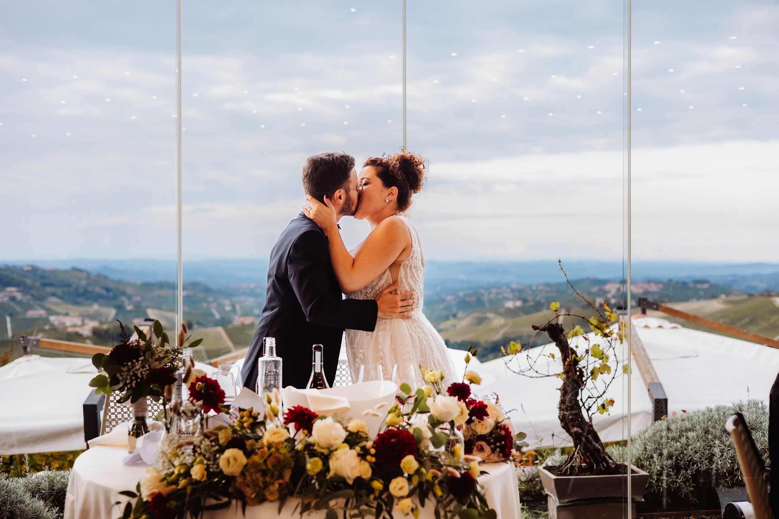 Couple shares romantic kiss at scenic mountain overlook during sunset wedding celebration with floral decor.