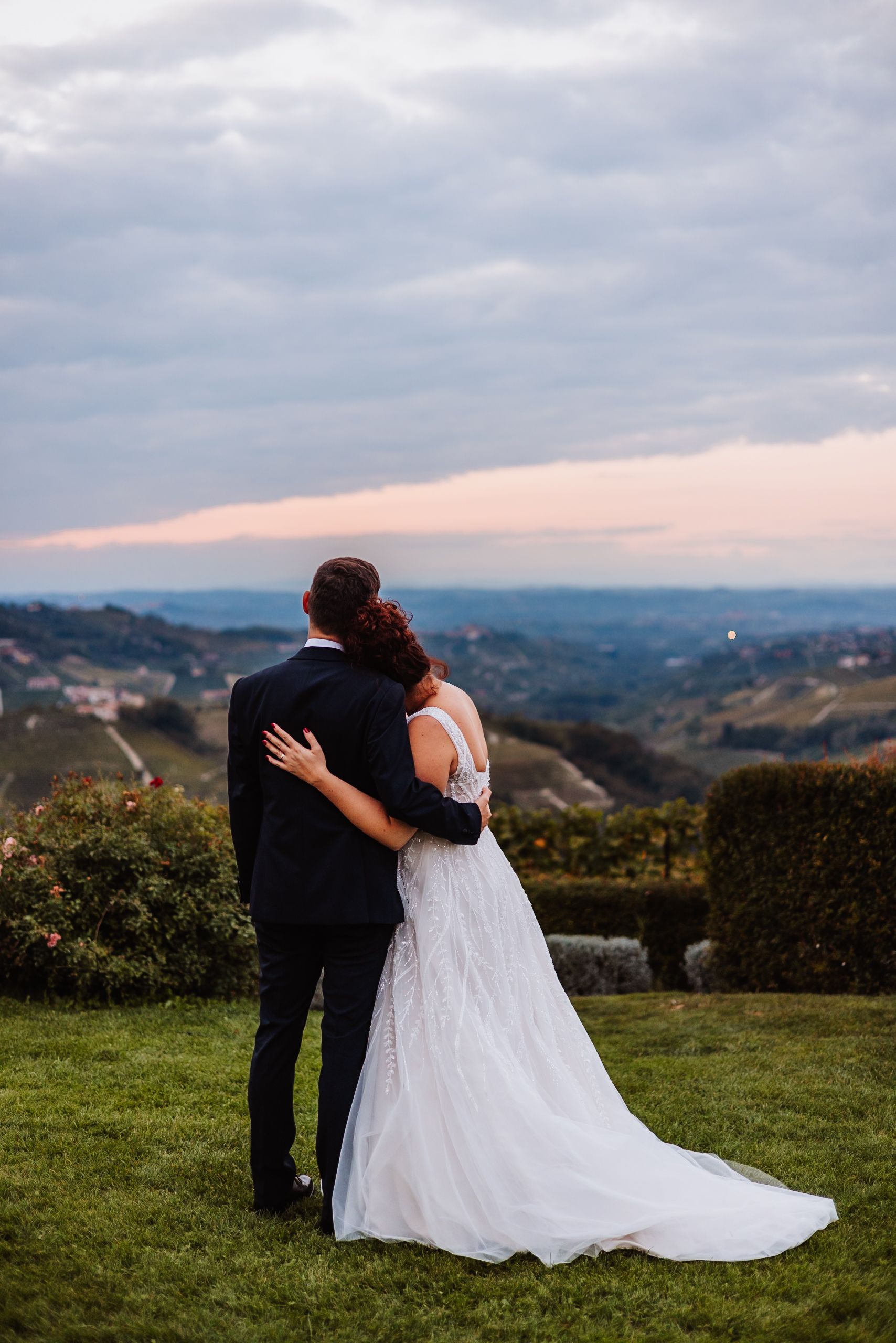 A couple embraces while overlooking a scenic mountain vista at sunset, the bride wearing a flowing white wedding gown.