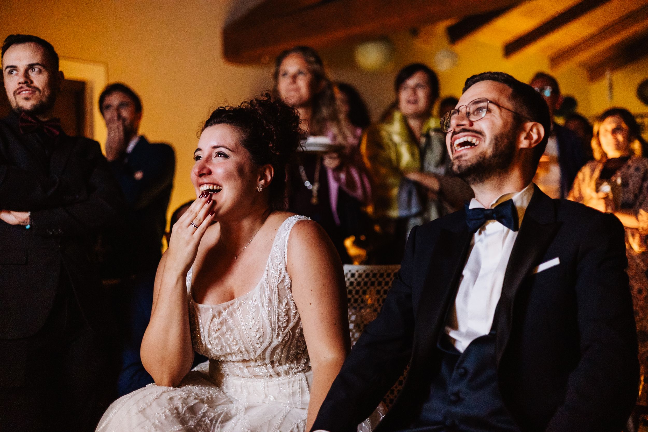 Wedding guests laugh joyfully while watching entertainment at an indoor reception venue with warm lighting.