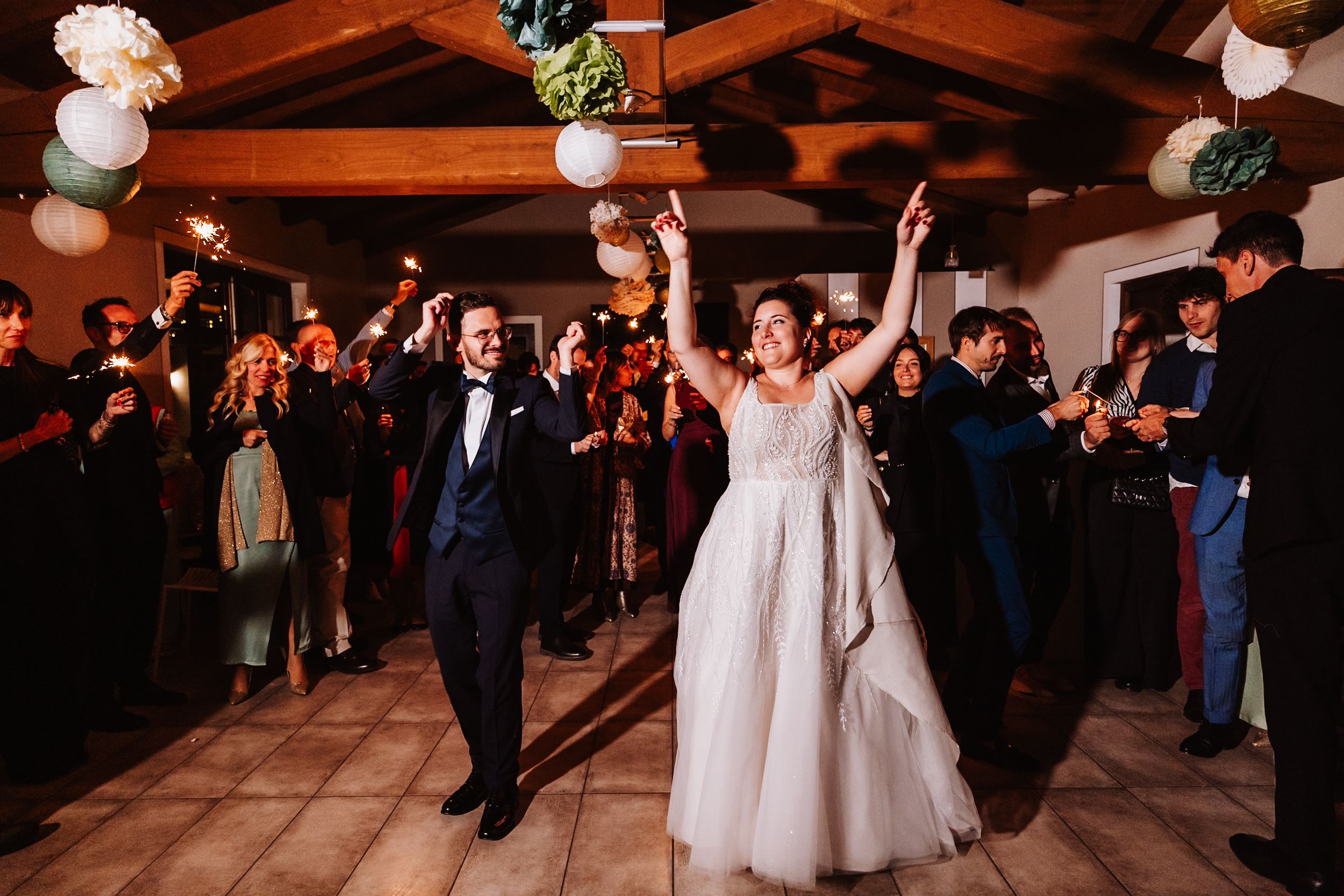 Newlyweds celebrate with guests on the dance floor under rustic wooden beams decorated with white floral arrangements.