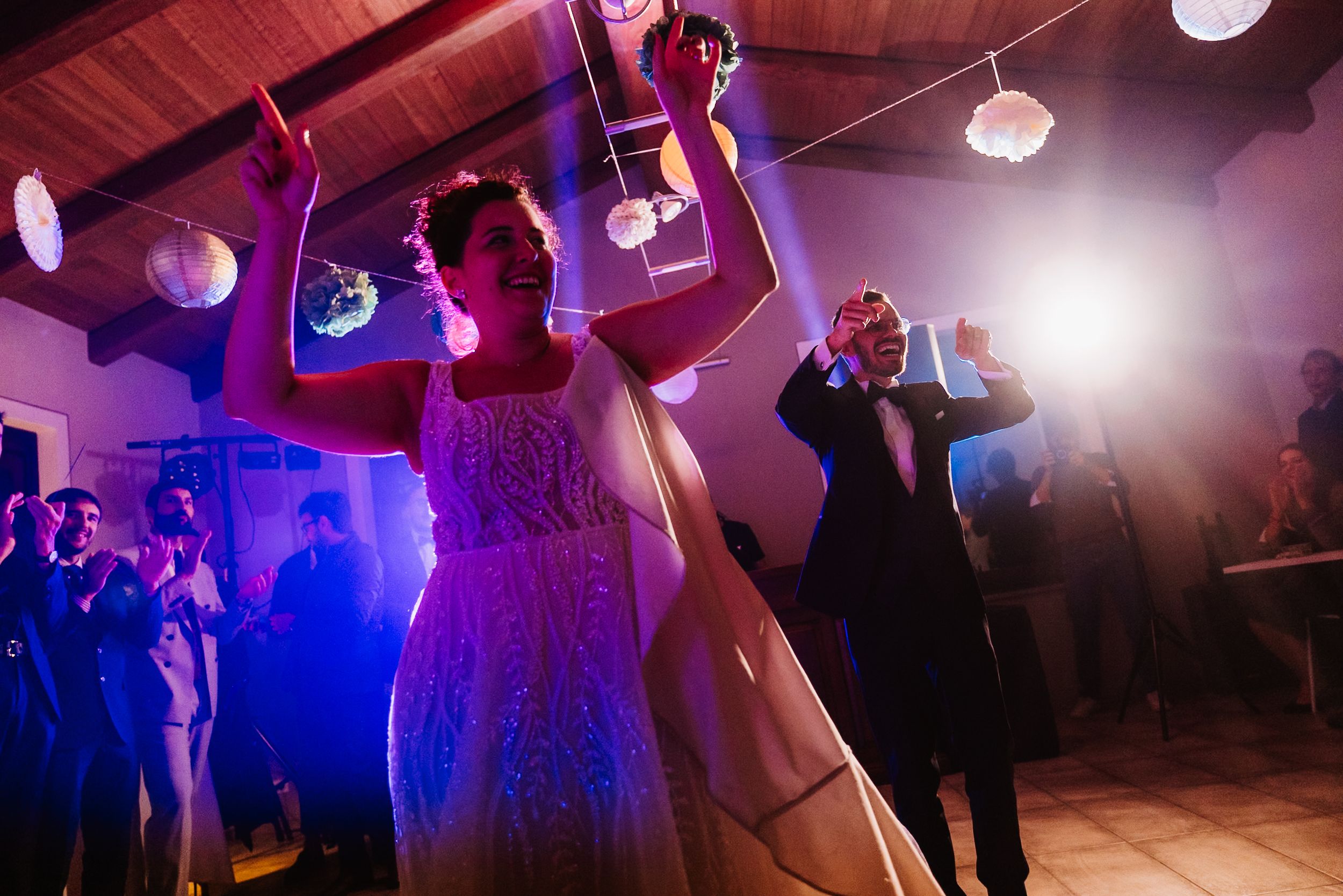 Wedding guests dancing and celebrating on a dance floor illuminated by colorful purple and blue lights at a reception.