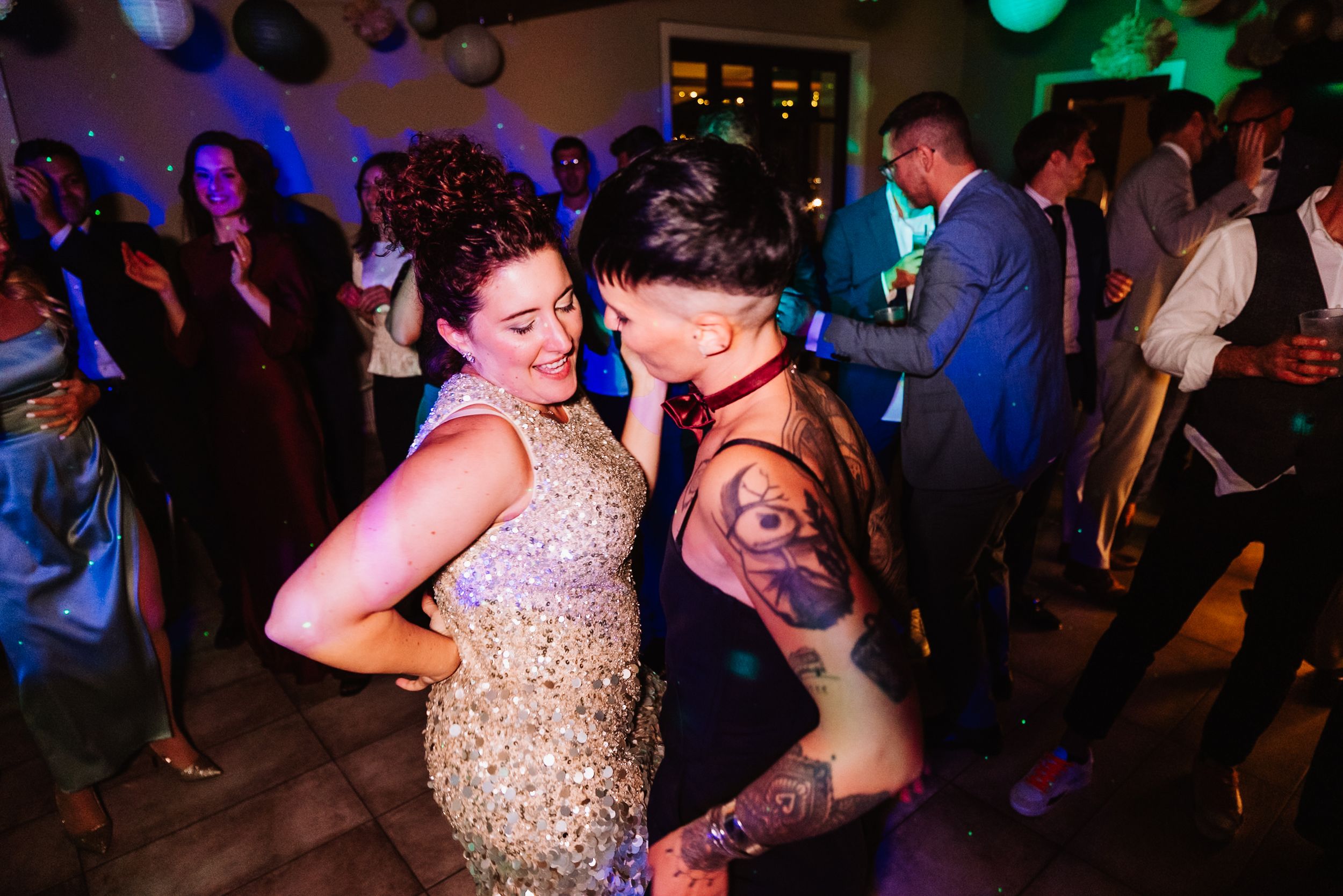 Wedding guests dance together at a reception under colorful party lights on the dance floor.