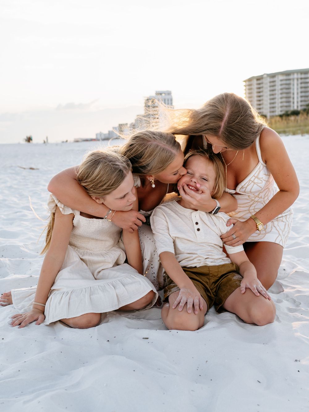 Family members lying together playfully on white sand beach wearing coordinated white outfits.