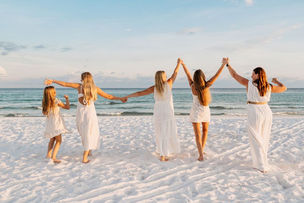 A group of friends in white dresses hold hands and dance together on a pristine beach at sunset.