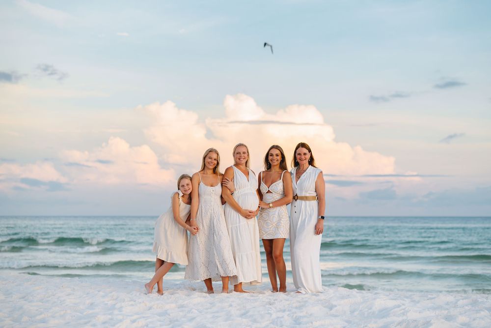 People in white outfits standing together on a beach with turquoise water and fluffy clouds at sunset.