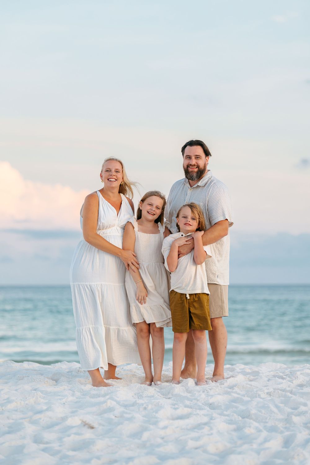Family portrait on a white sandy beach with ocean backdrop wearing coordinated white and khaki outfits.