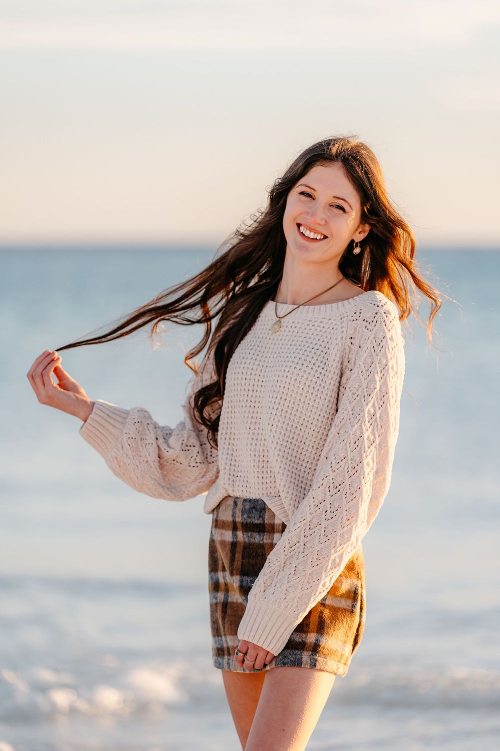 Young woman in cream knit sweater and plaid shorts laughs joyfully at sunset on a beach.