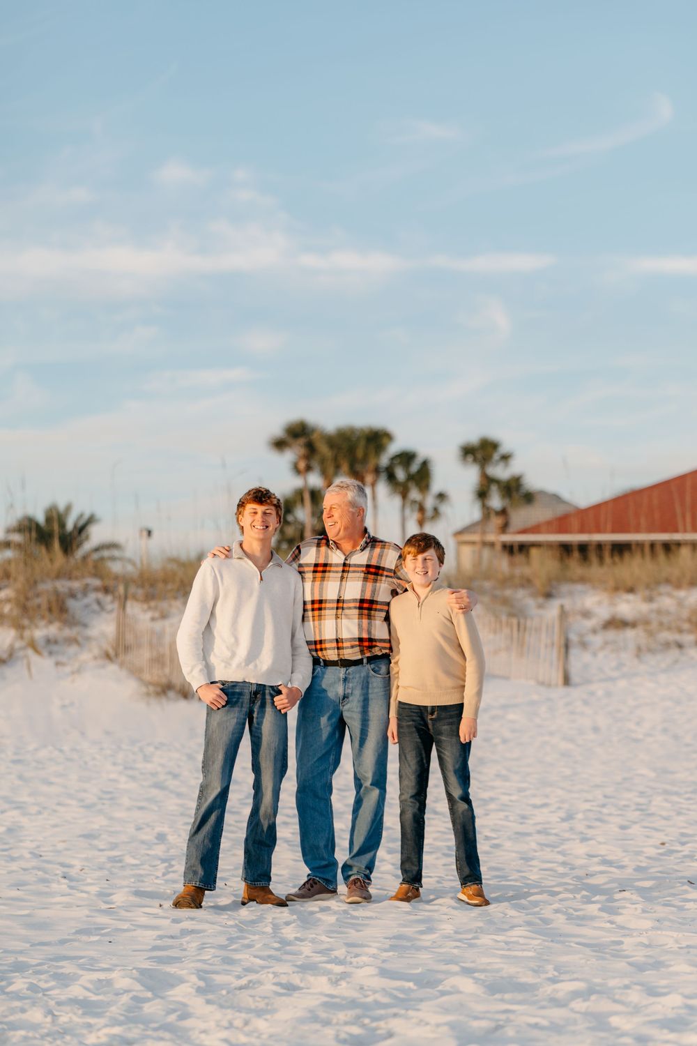 Three people standing together on white sandy beach during sunset with palm trees in background.