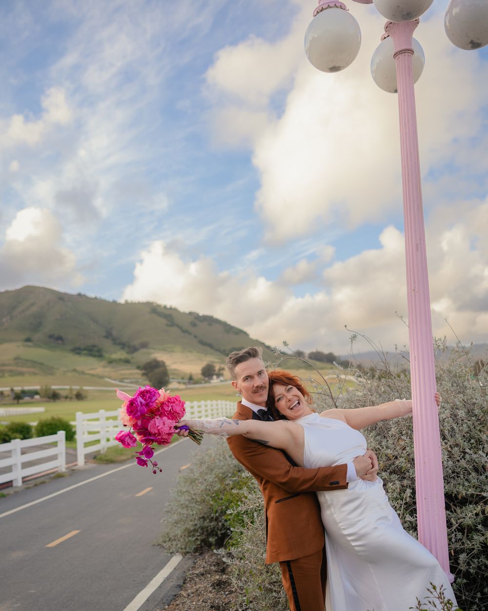 A couple poses playfully in a series of photos along a scenic mountain road with white fencing and pink flowers.