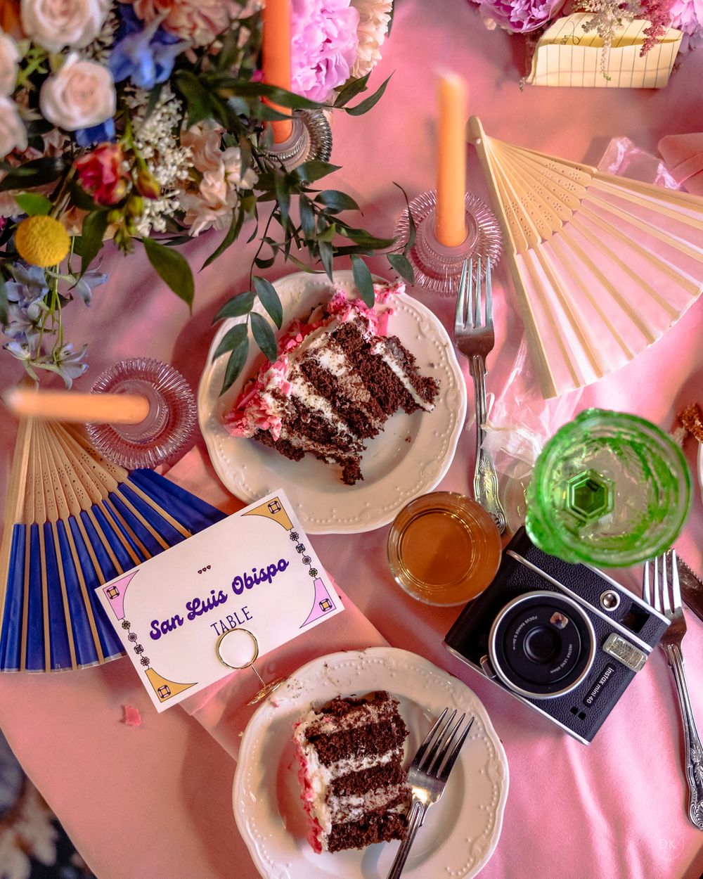 Chocolate layer cake slices with pink frosting on plates alongside vintage camera, paper fans and colorful drinks on pink table.