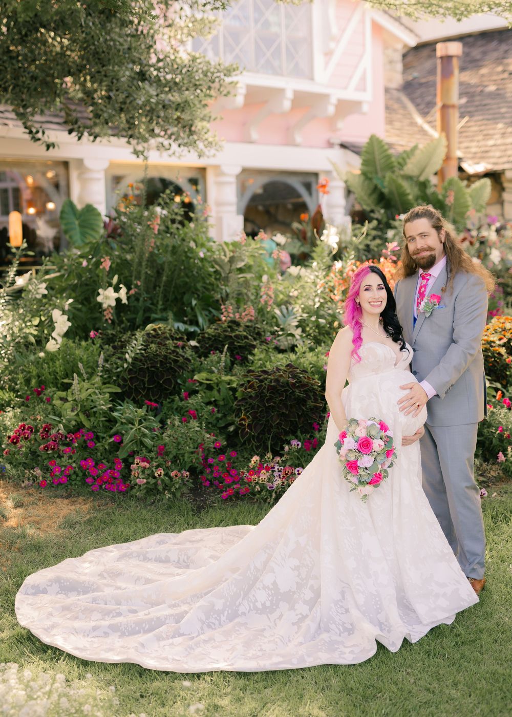 A romantic wedding couple poses in a garden setting with pink flowers and a flowing white lace gown.
