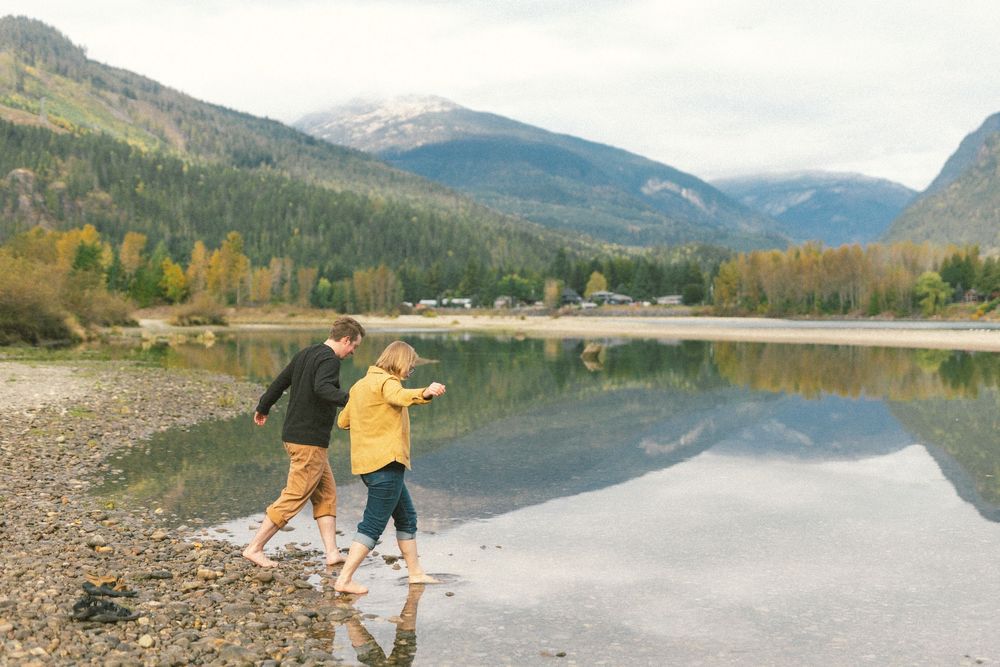 lesbian couple wade into the Columbia river with fall colours on the mountains behind them in Revelstoke, BC