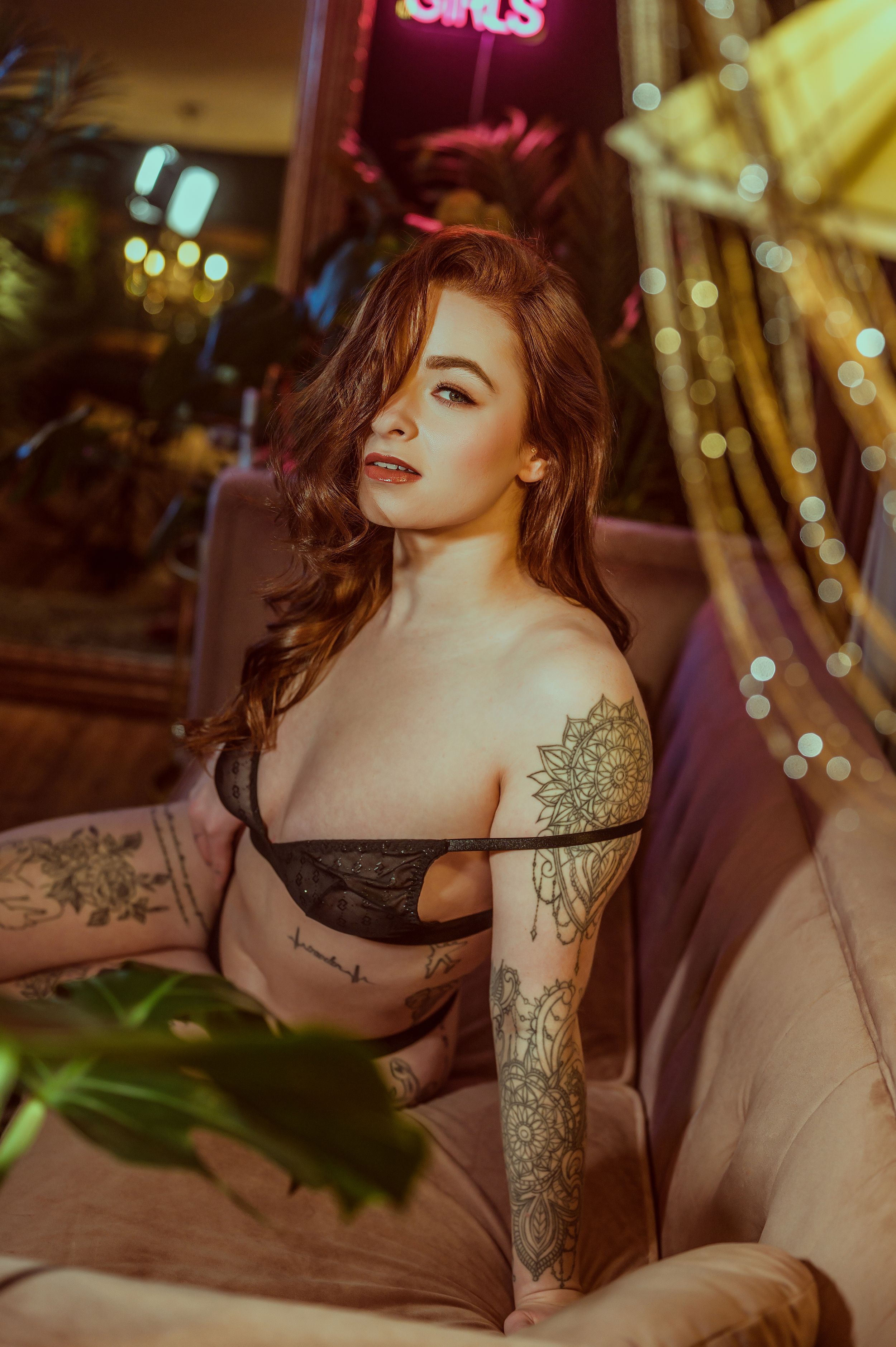 Artistic boudoir photoshoot with moody lighting features tattooed model on couch with bokeh lights in background.