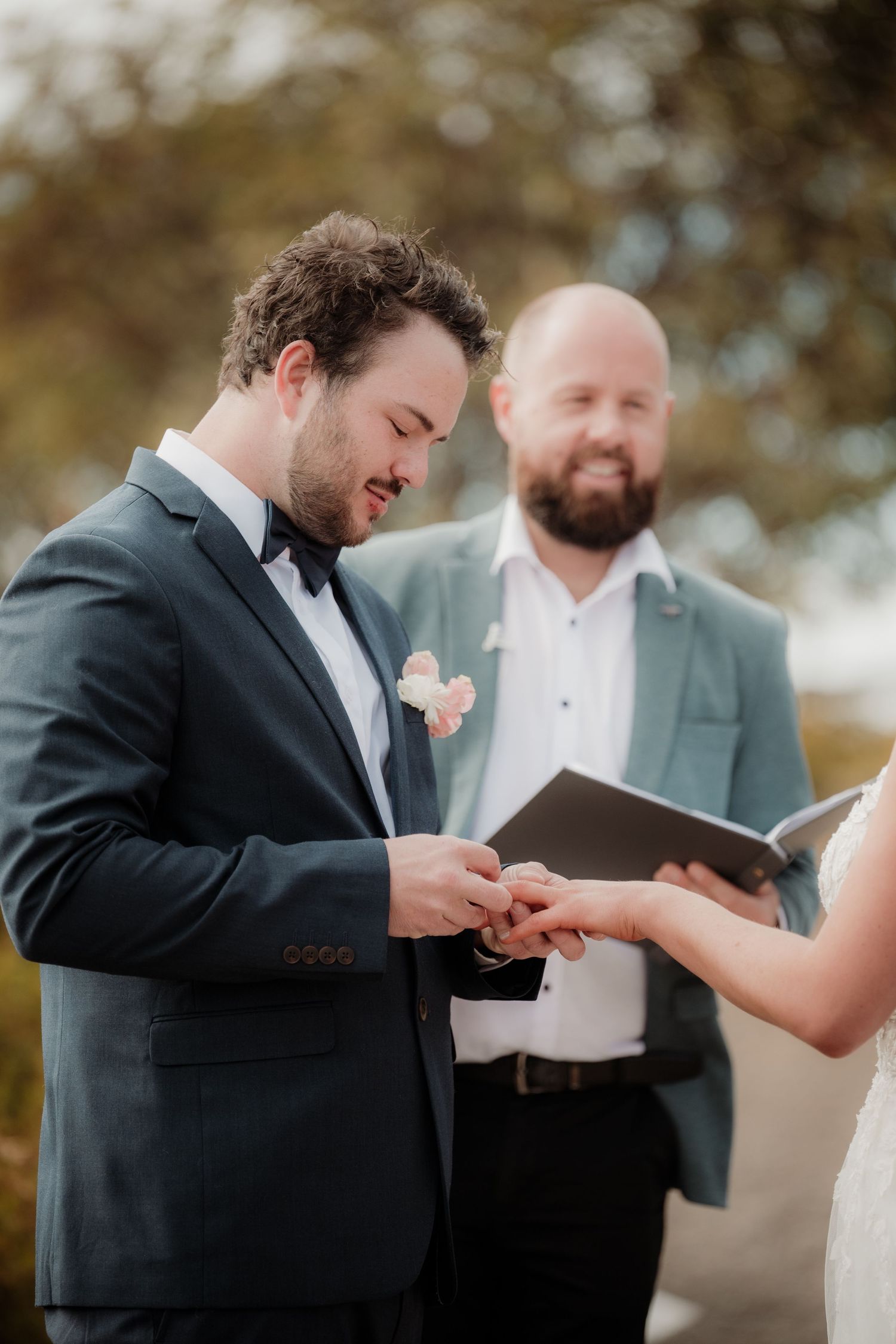 Groom and groomsman share a lighthearted moment during ring exchange at outdoor wedding ceremony.