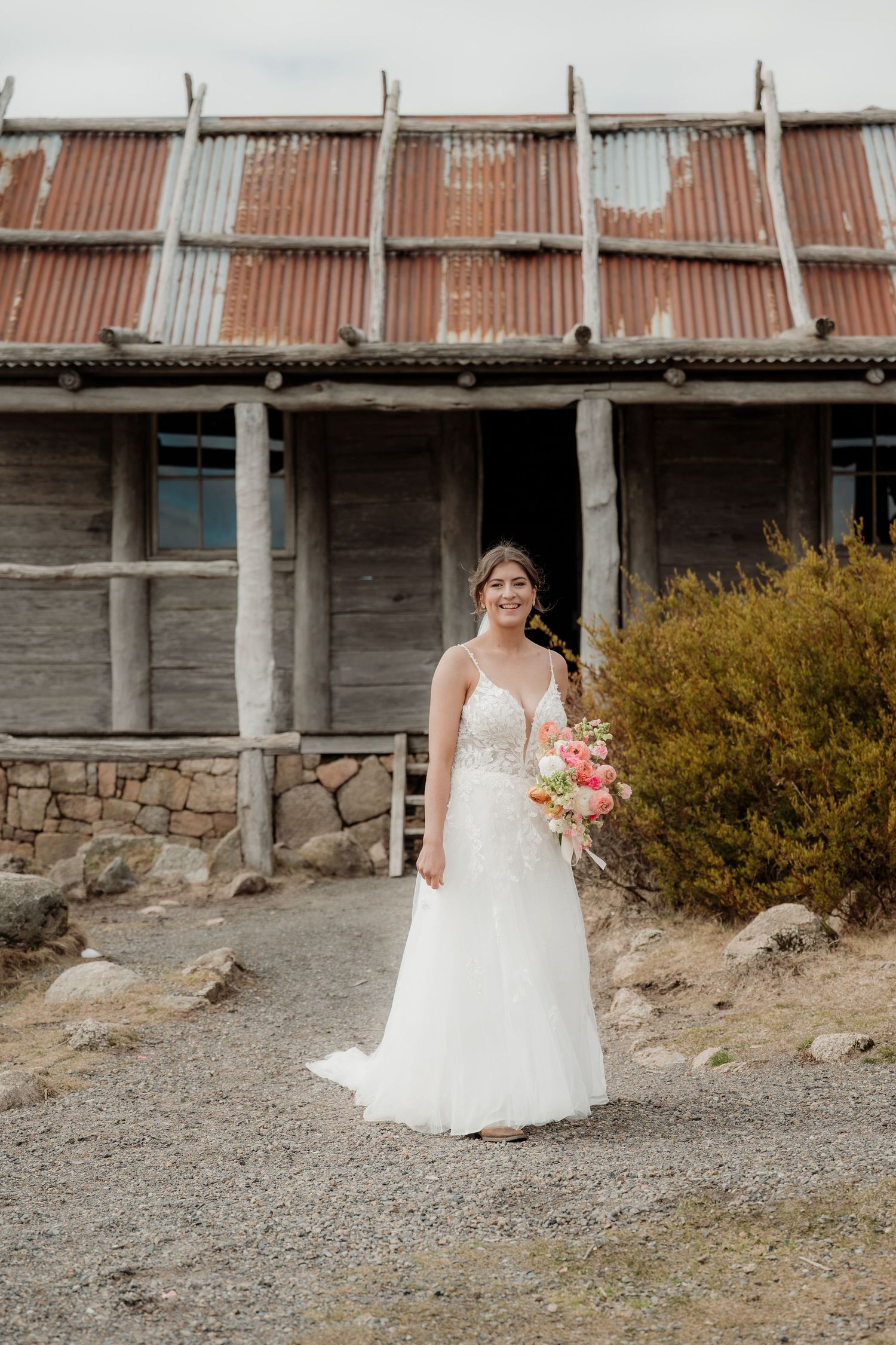 A person in a white wedding dress holds a bouquet while standing against a rustic barn wall and desert landscape.
