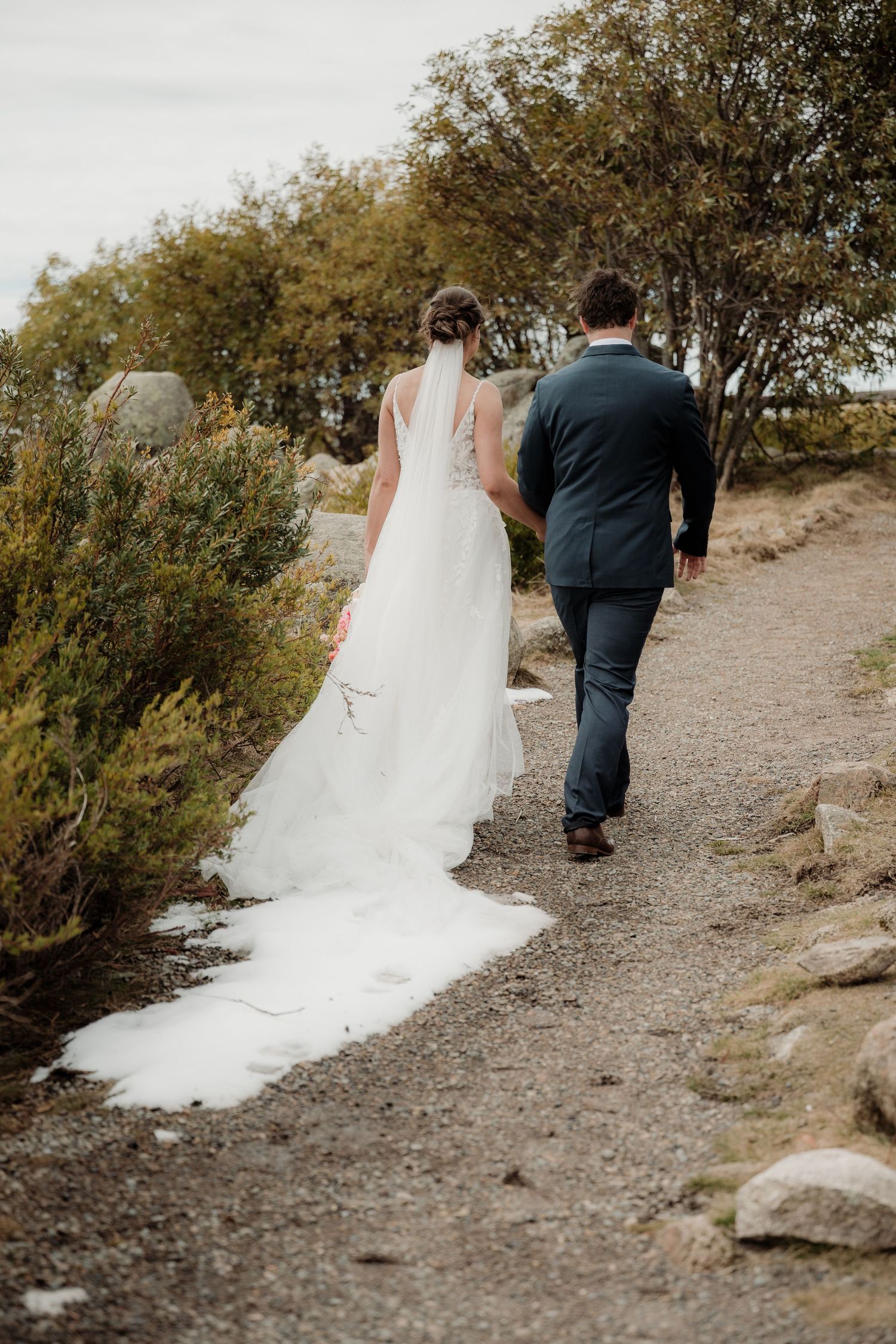 A couple in wedding attire walking down a rustic dirt path surrounded by autumn trees and rocks.