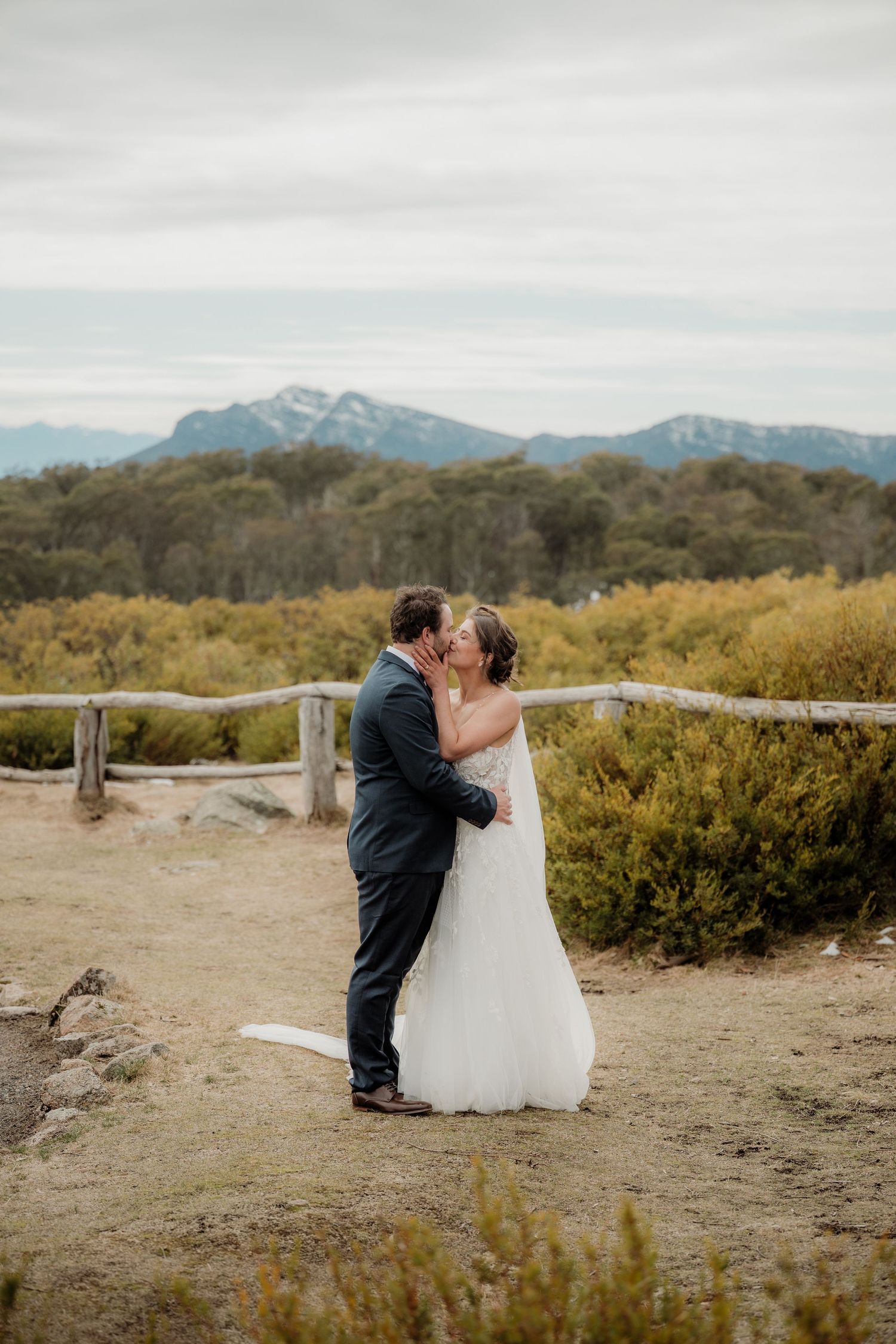 A couple shares a romantic moment on a mountain trail with scenic mountain ranges and brush in the background.
