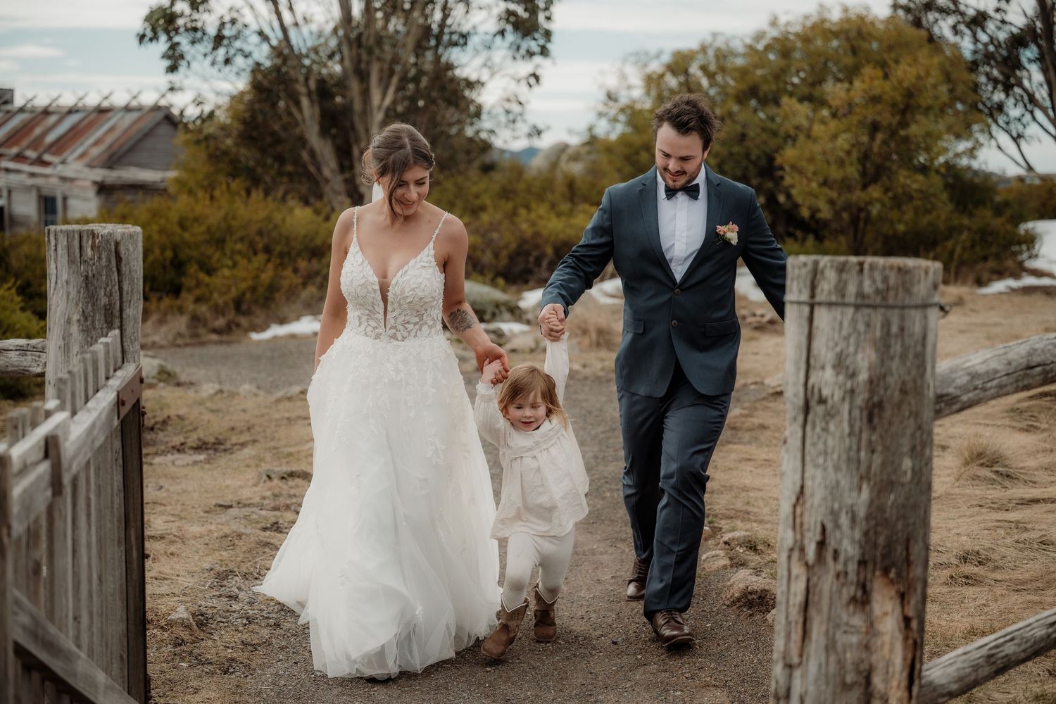 A couple walks hand in hand with a child along a rustic wooden fence at sunset in an outdoor setting.