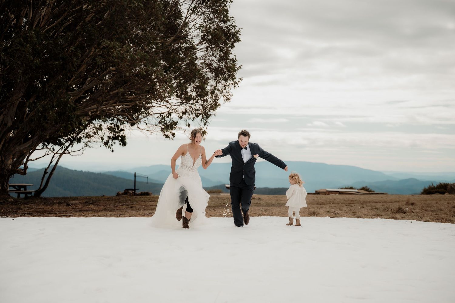 Three people joyfully running together on a snowy mountain path with scenic mountain views in the background.