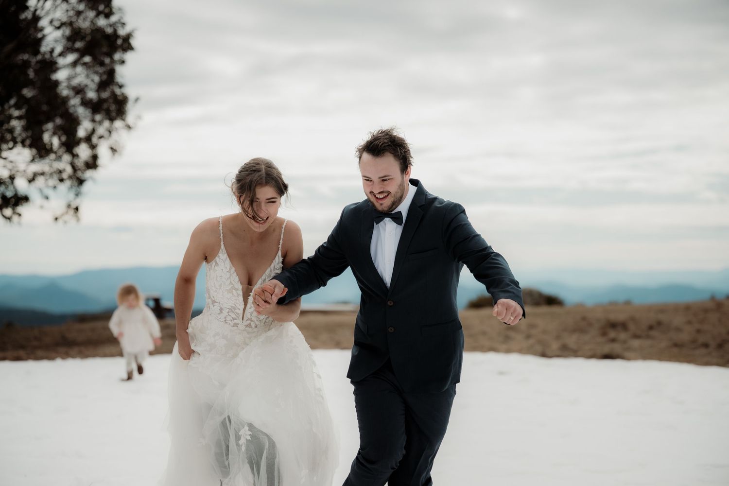 A couple in wedding attire joyfully running together on a hilltop against a cloudy mountain landscape.