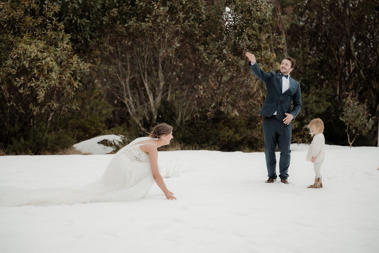 A couple in winter wedding attire playfully throws snow at each other in a snowy outdoor setting.