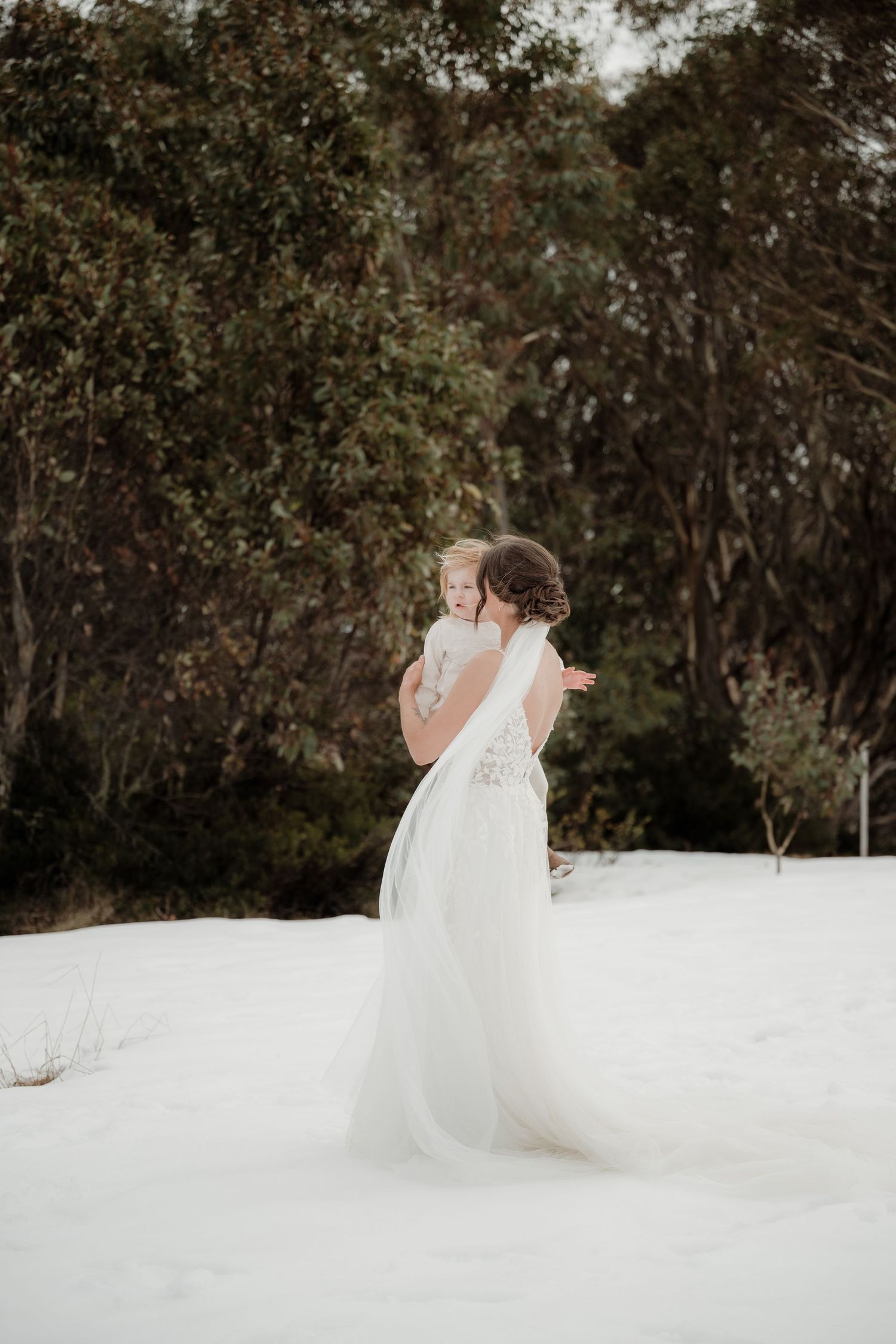 A bride in a flowing white wedding dress twirls gracefully in a natural outdoor setting among trees.