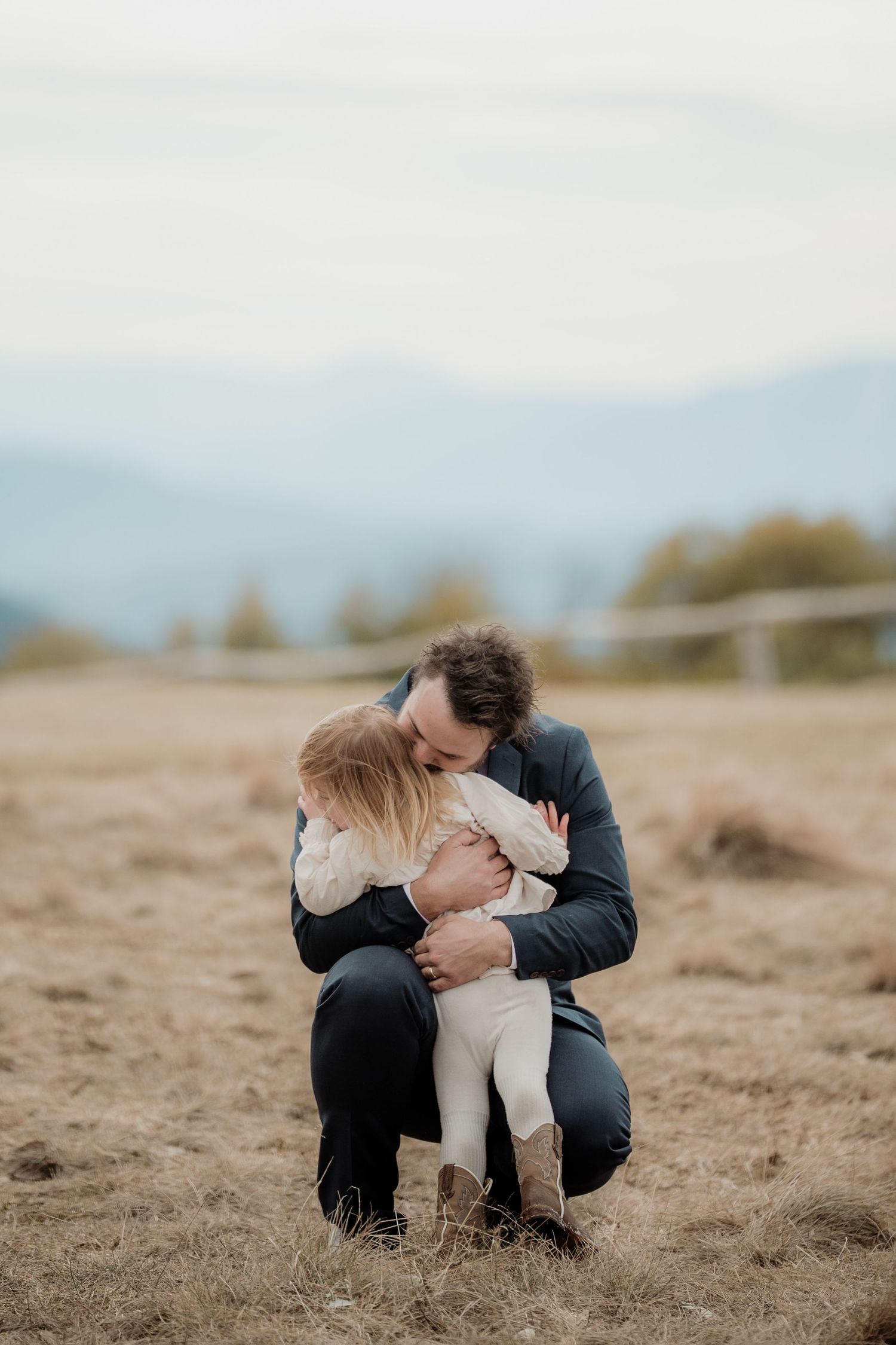 A couple shares a tender moment while sitting together on a sandy beach with mountains in the background.