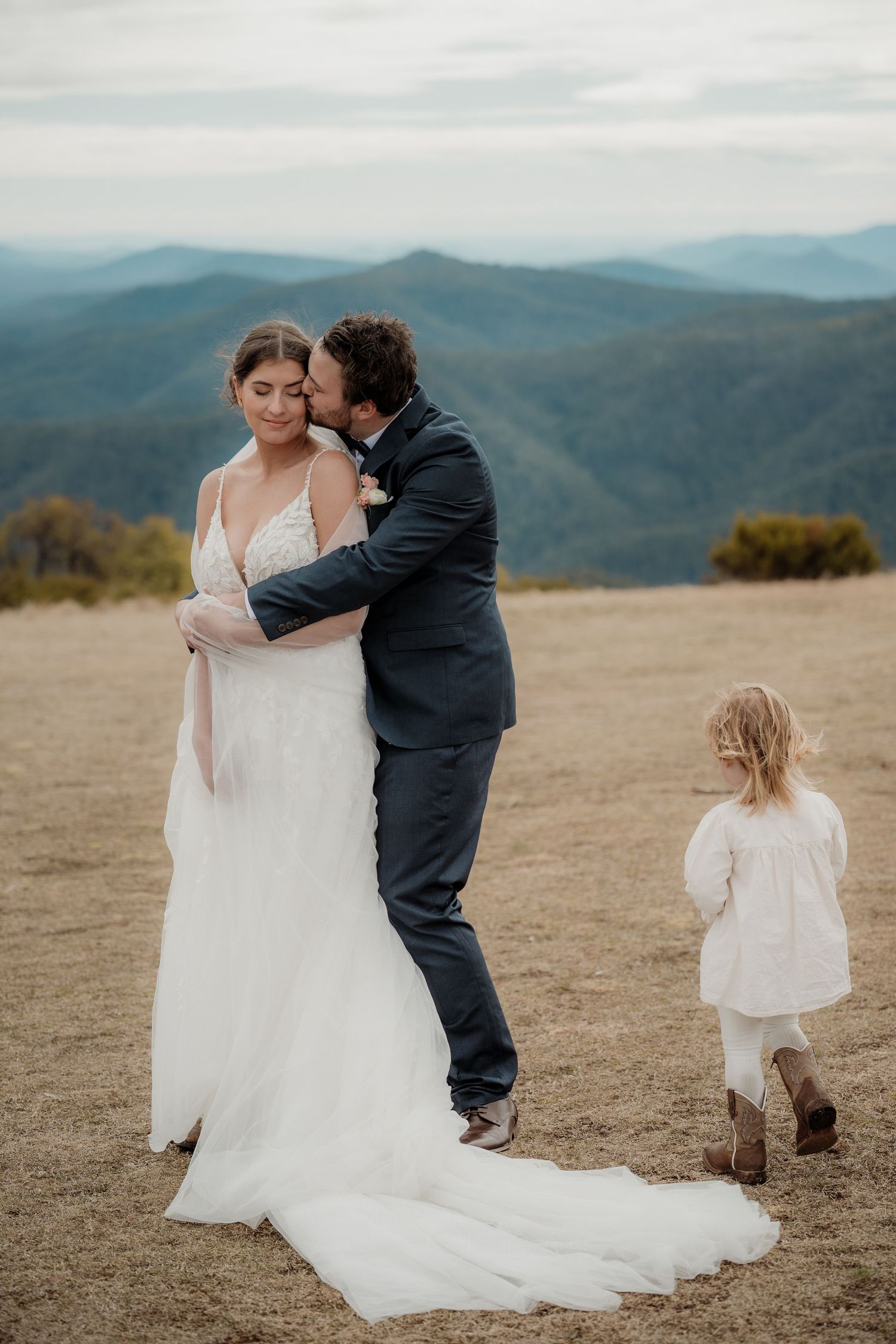 A couple shares an intimate moment on a mountaintop with scenic mountain ranges in the background.