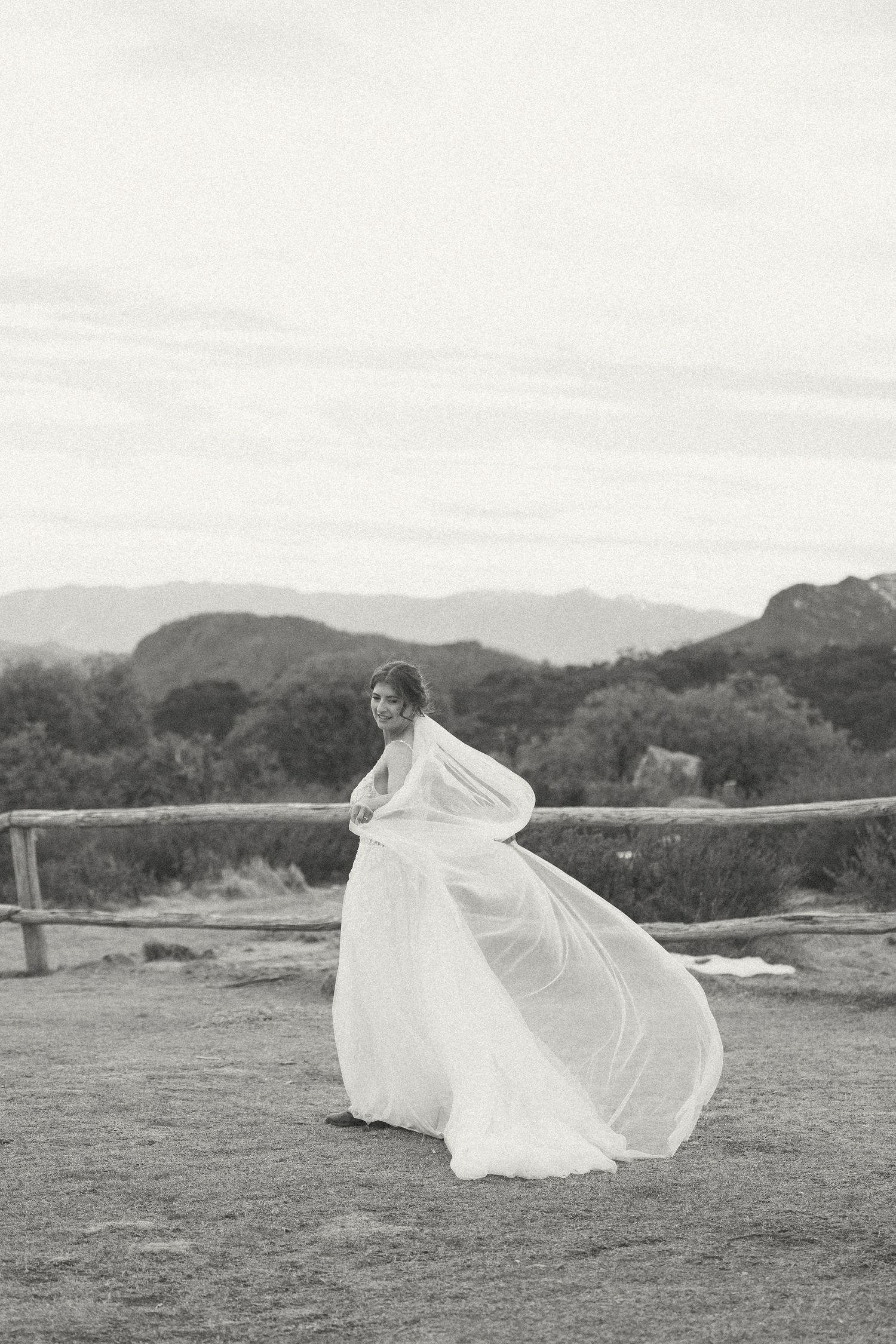 A flowing white wedding veil billows dramatically in the wind across a grassy hillside landscape.
