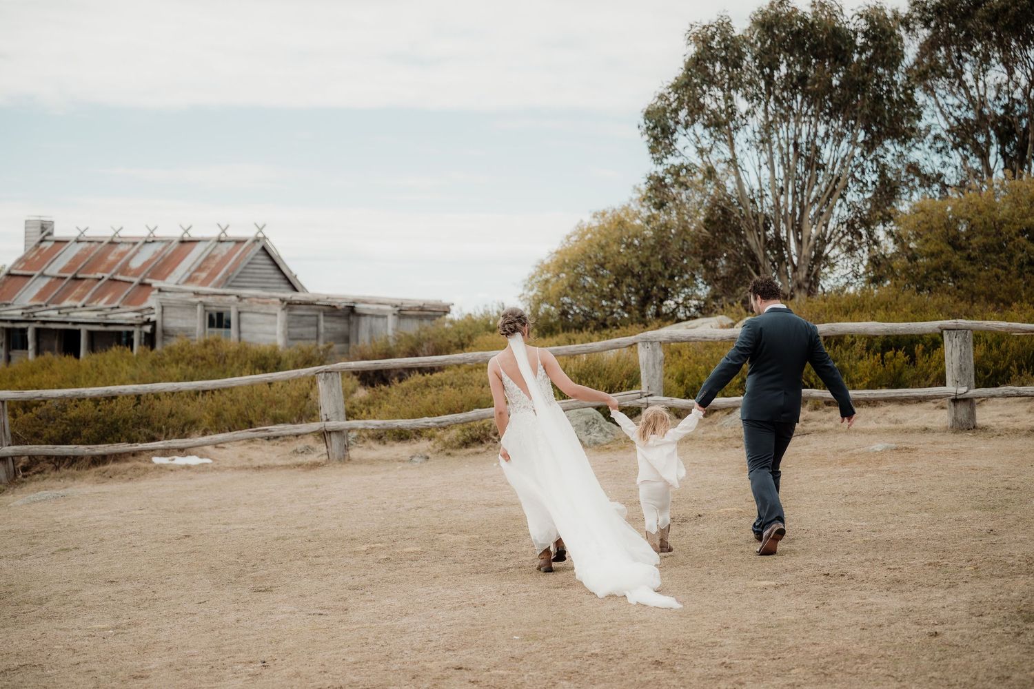 A couple in wedding attire runs playfully along a rustic wooden fence with a weathered barn in the background.