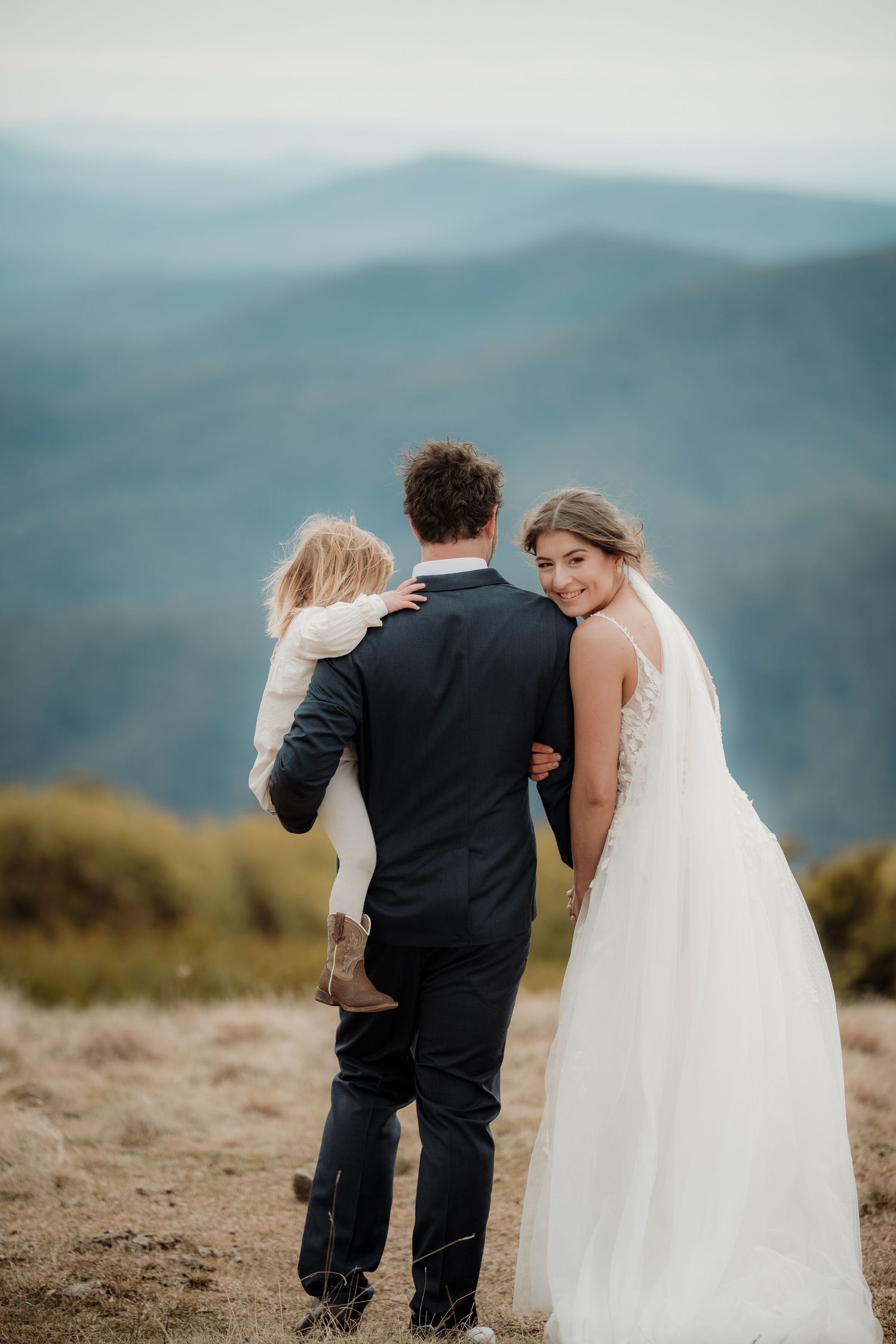 A couple in wedding attire stands on a mountaintop overlooking misty mountain ranges in the distance.
