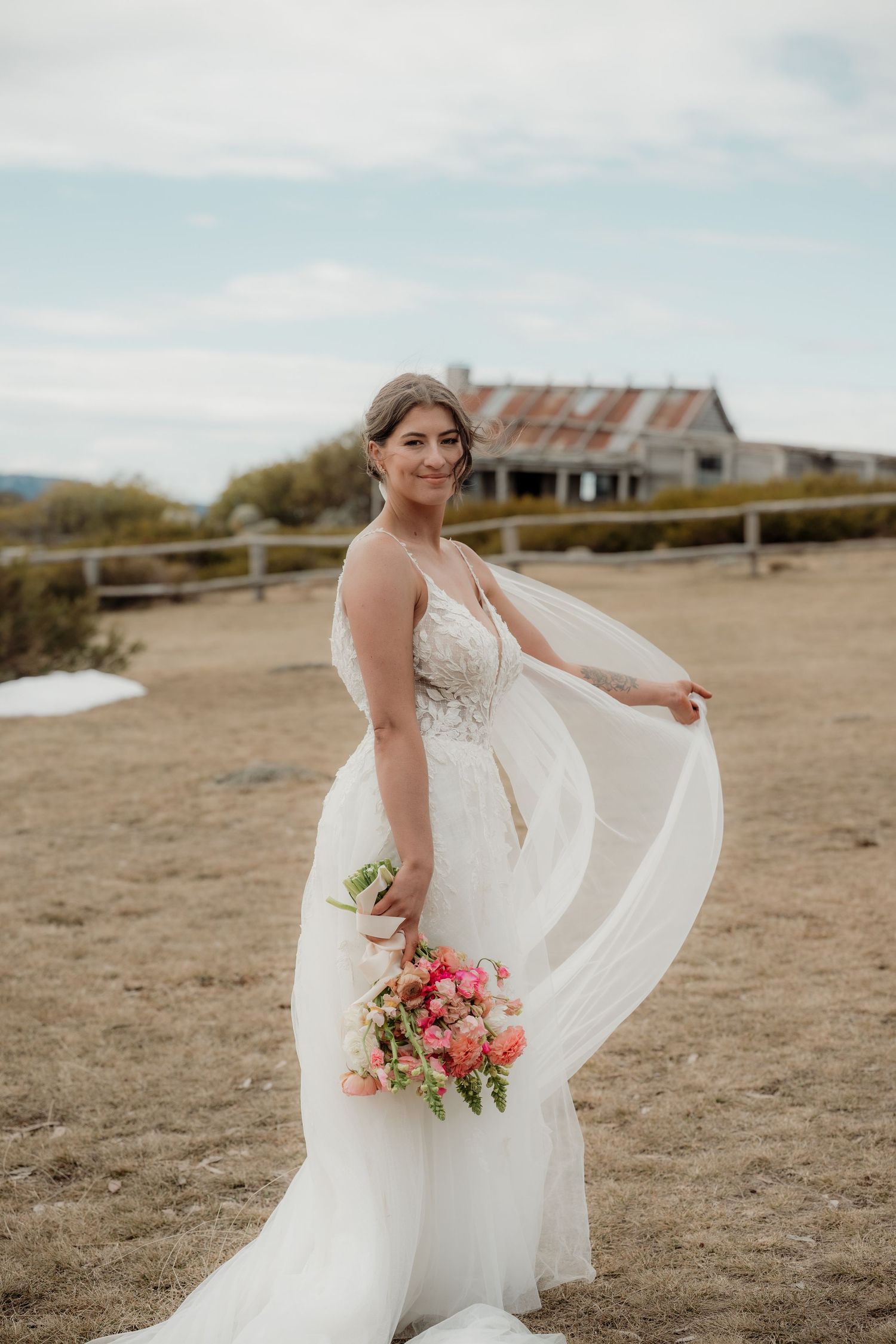 A bride in a flowing white dress holds a pink and coral bouquet while posing in an outdoor field setting.