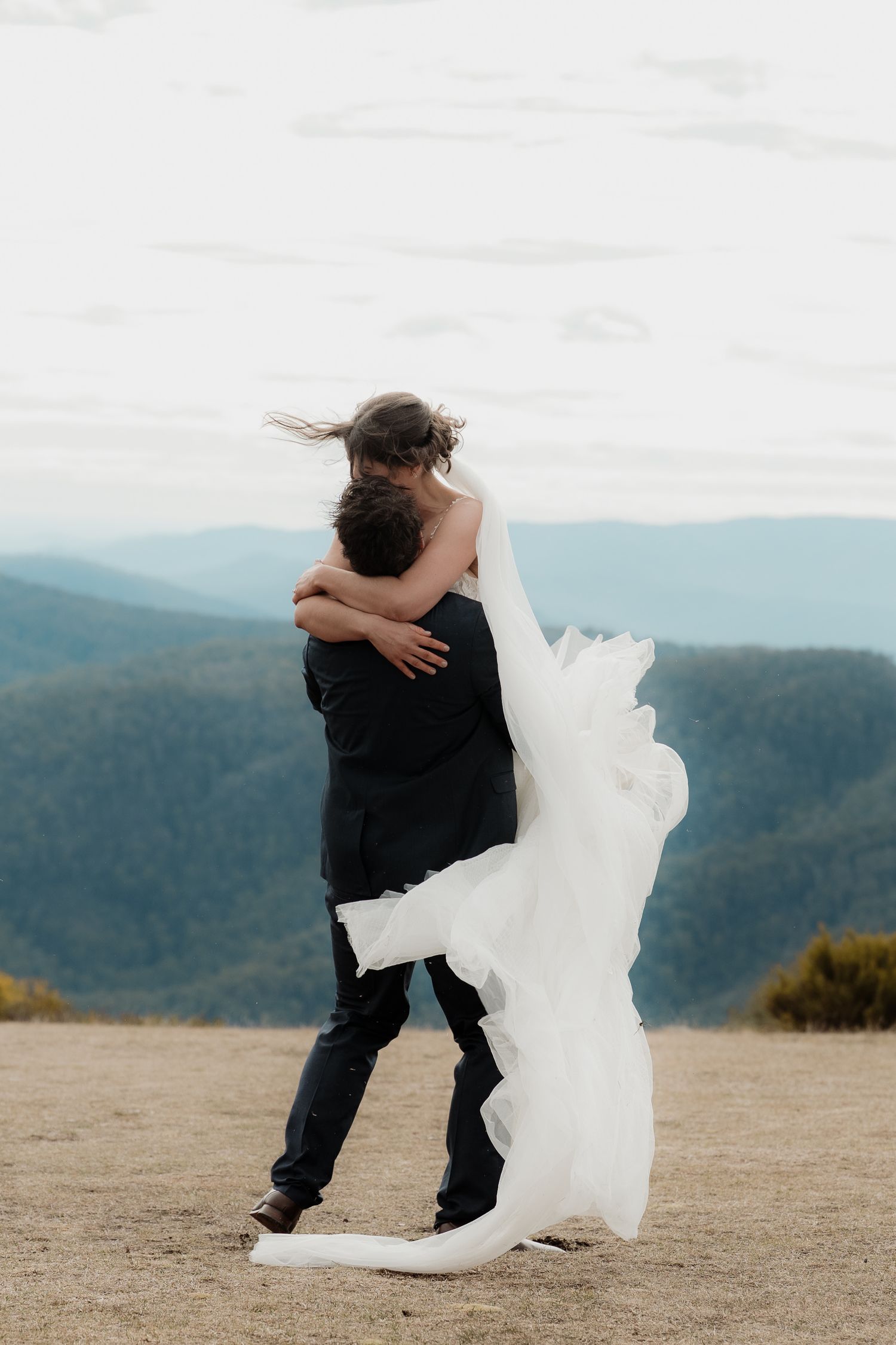 A person in black clothing playfully dances with a large fluffy white pillow against a mountainous landscape backdrop.