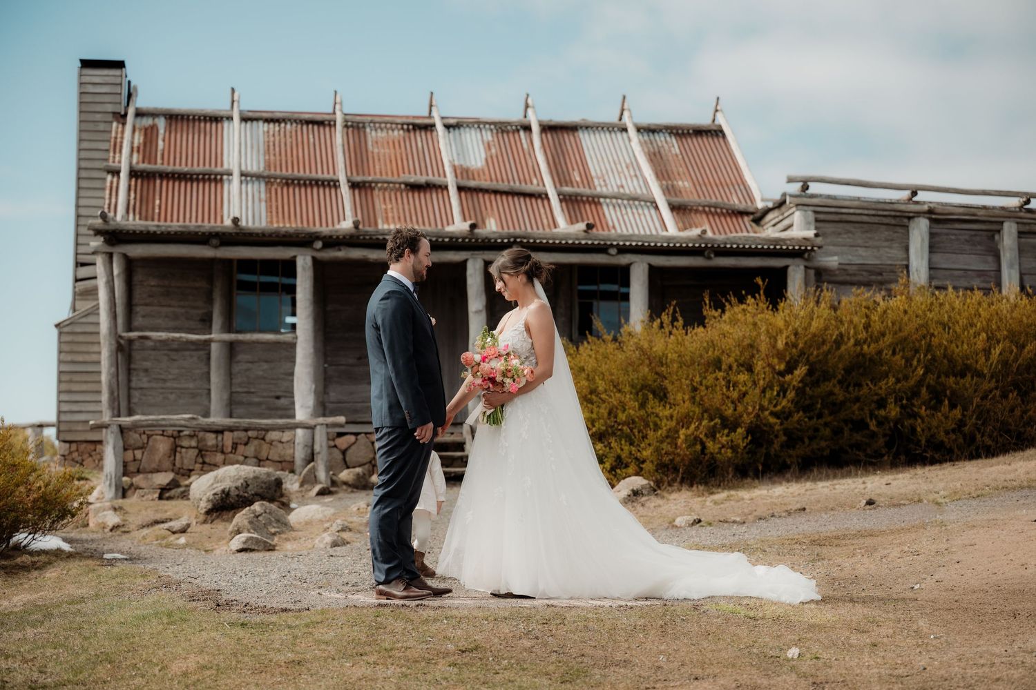A couple in wedding attire walks along a rustic beachside path with weathered buildings in the background.