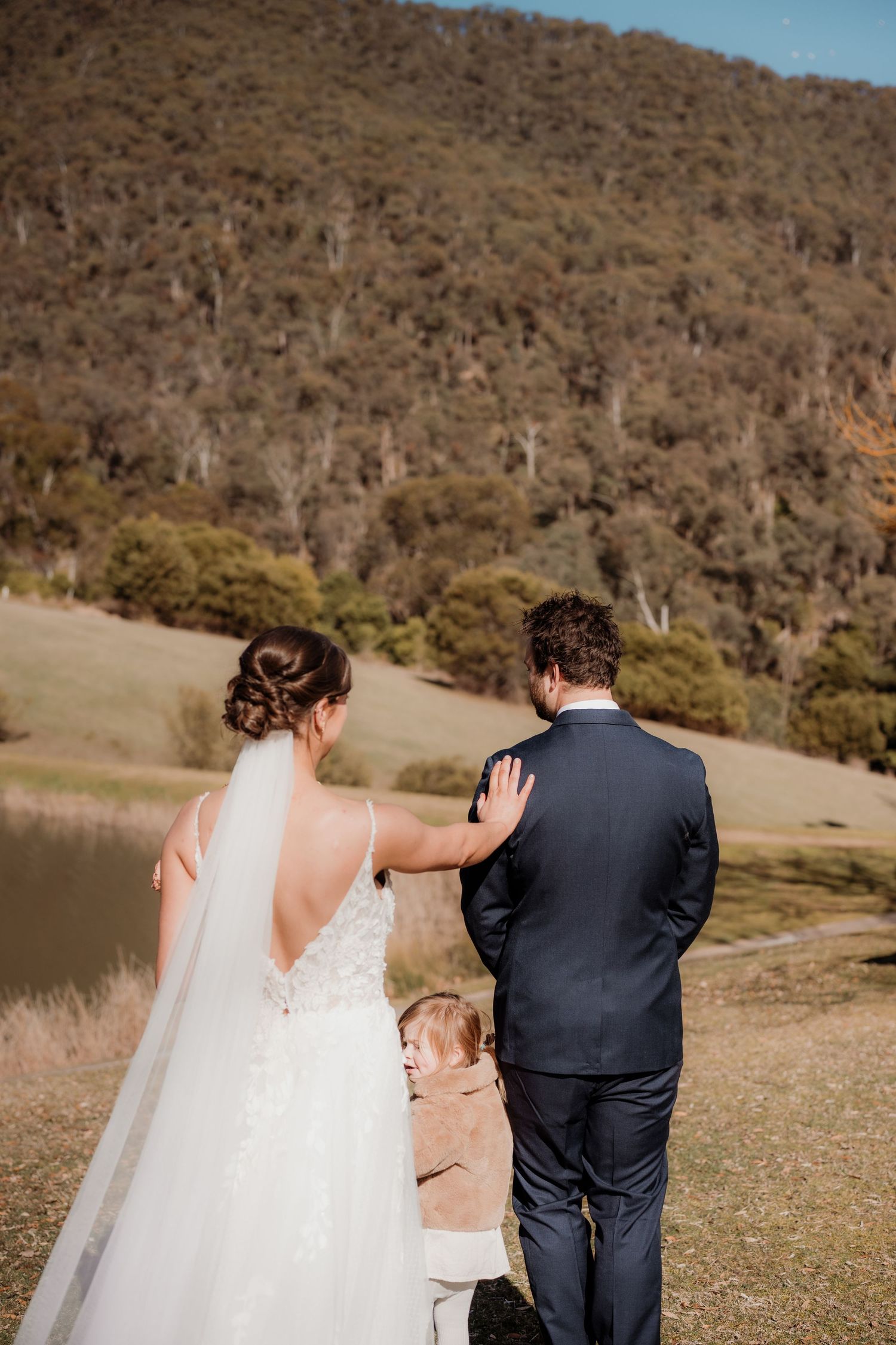 A couple stands together on a grassy hillside overlooking a serene lake with mountains in the background.